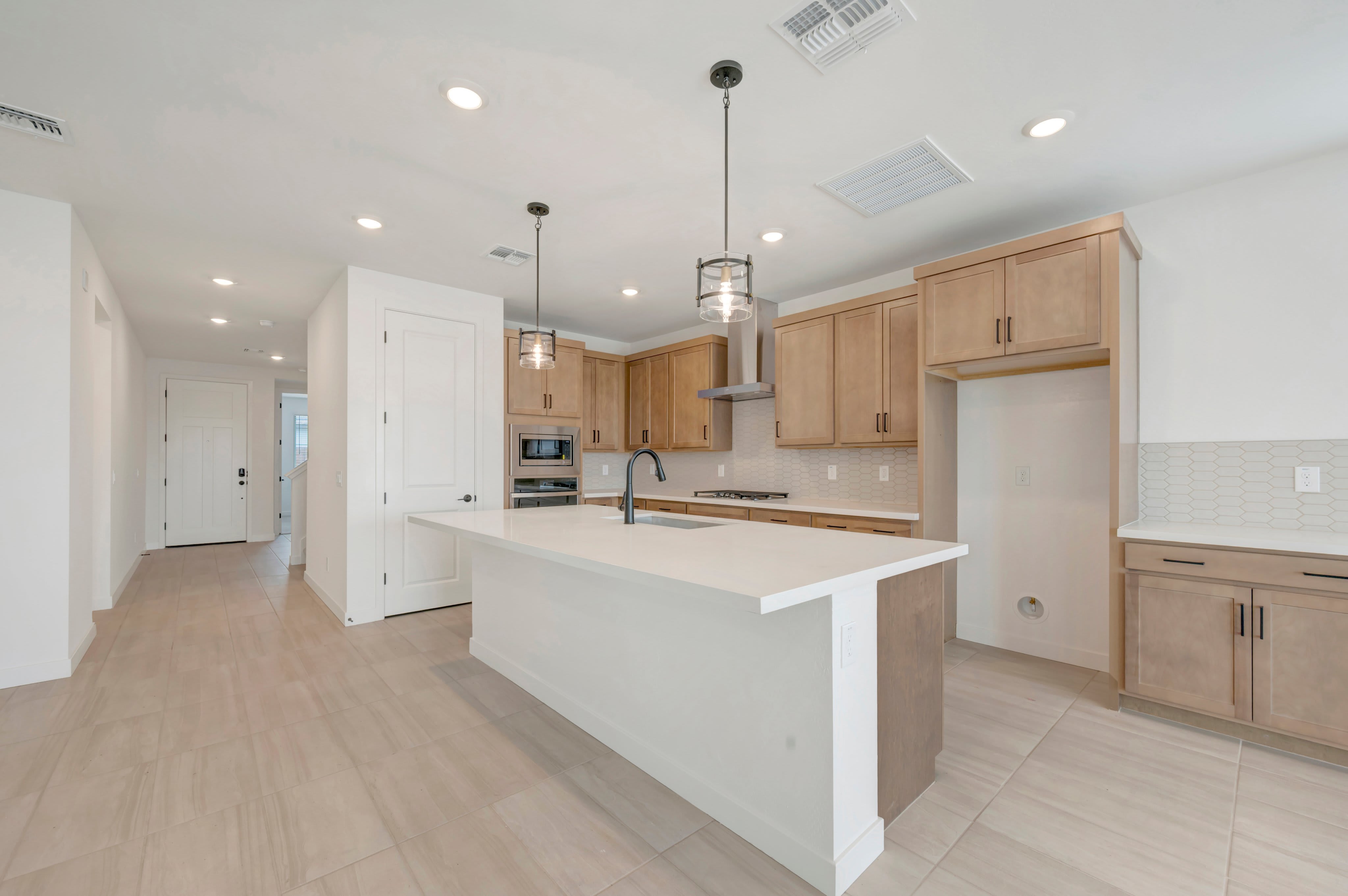 A large white kitchen island sits in the foreground of a modern kitchen featuring light wood cabinetry and a white tiled backsplash.