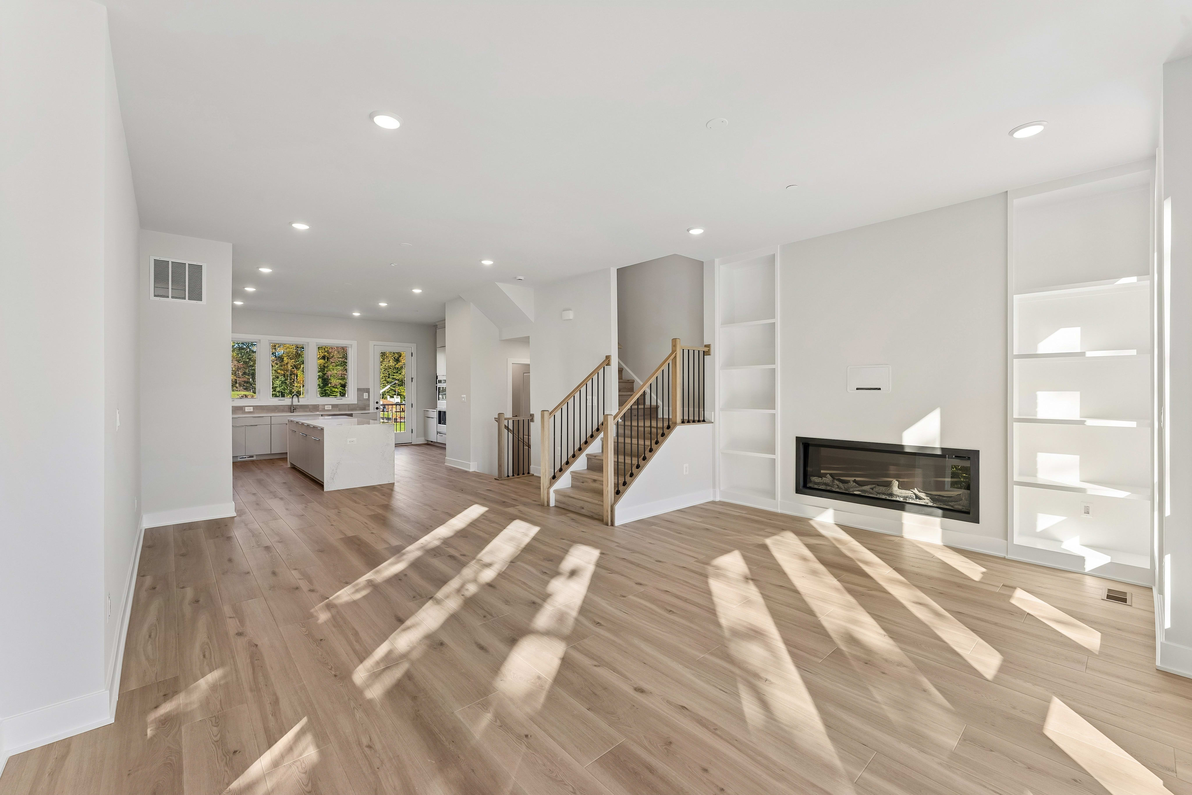 Sunlight streams across a light wood floor in an open living area, with a fireplace and built-in shelves in the foreground and a kitchen and staircase in the background.