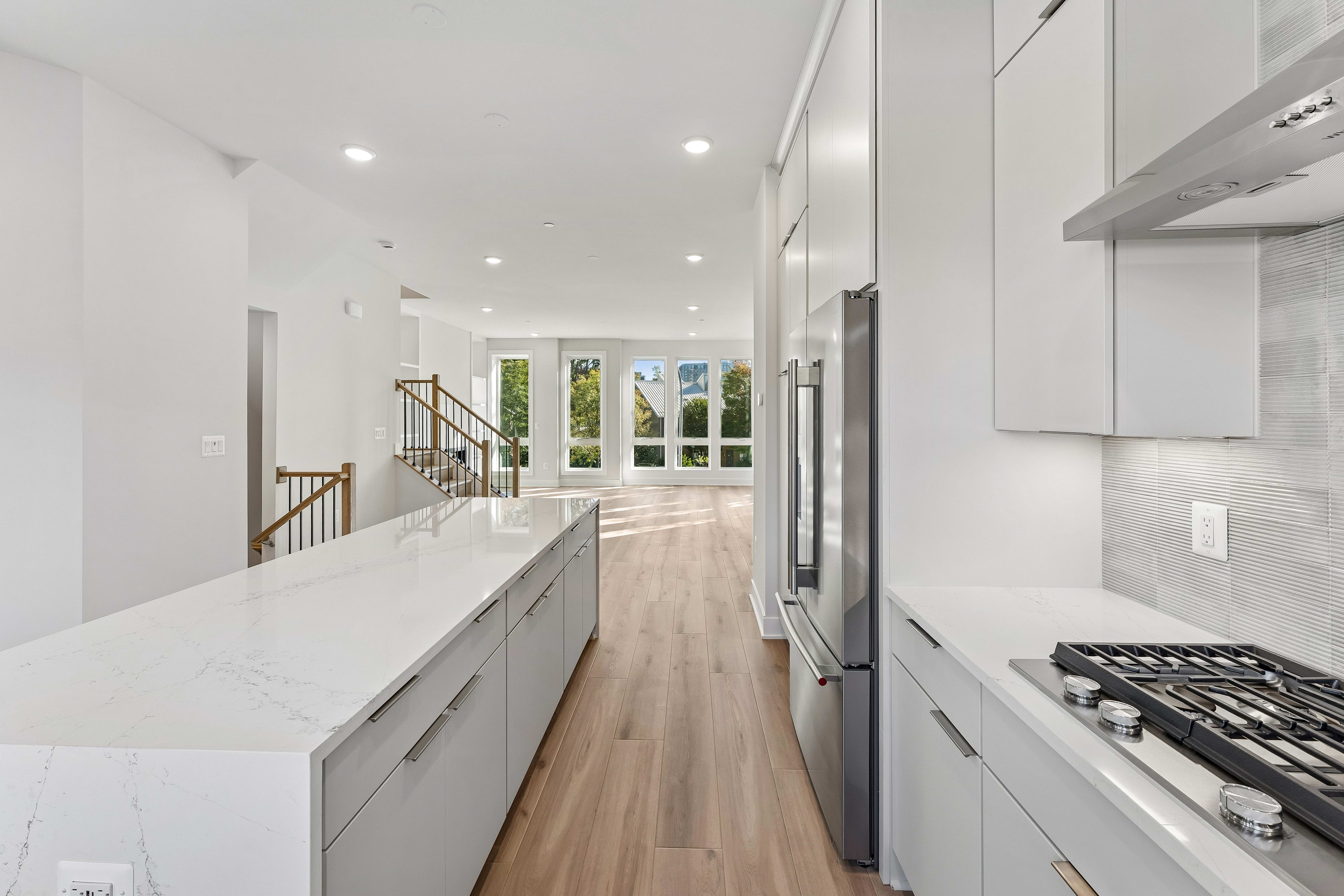 A modern white kitchen with a long marble island and stainless steel appliances leads toward a bright living area with wooden stairs and glass doors.