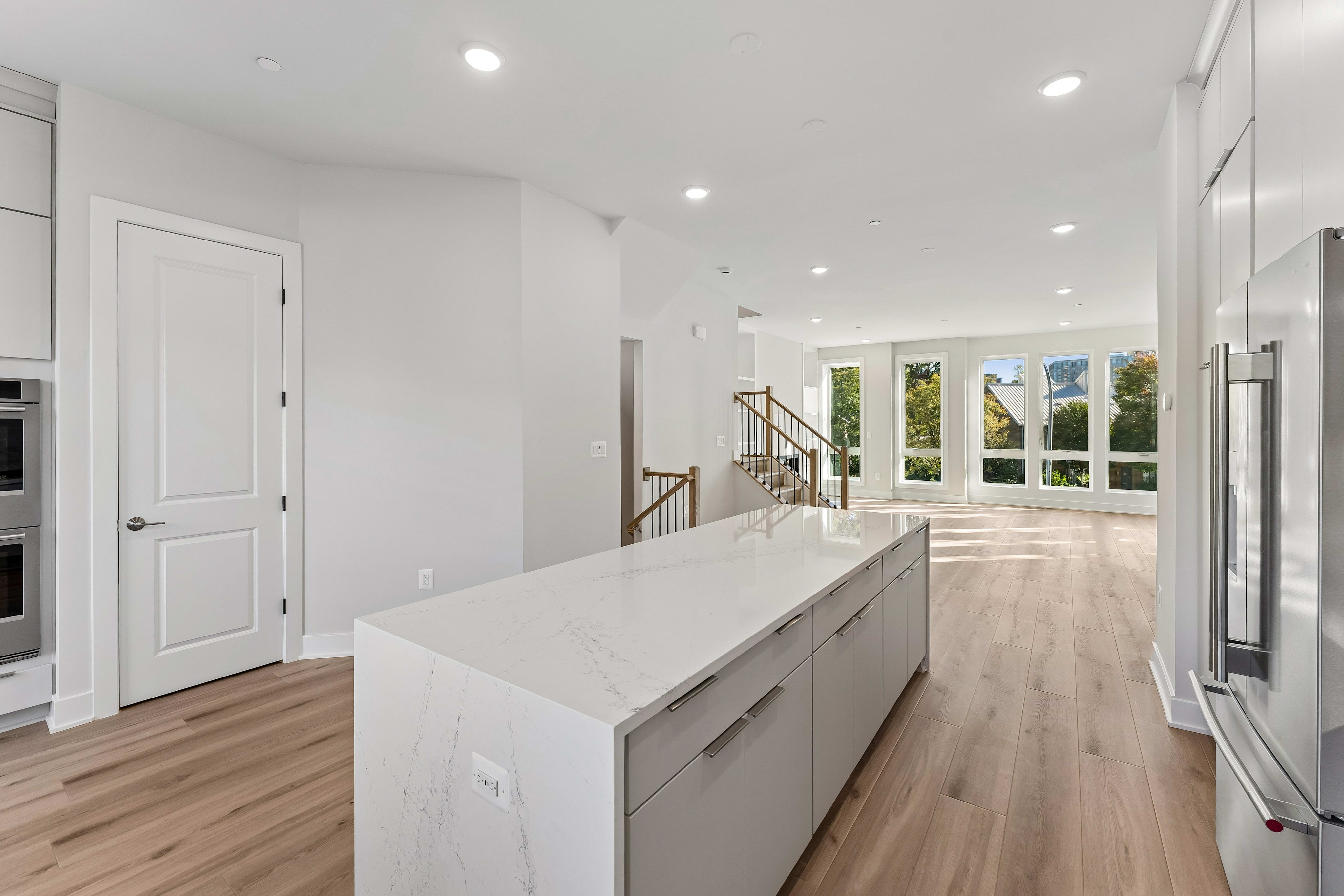 A white marble kitchen island sits in the foreground of a bright, open-concept room with light wood floors and large windows in the background.