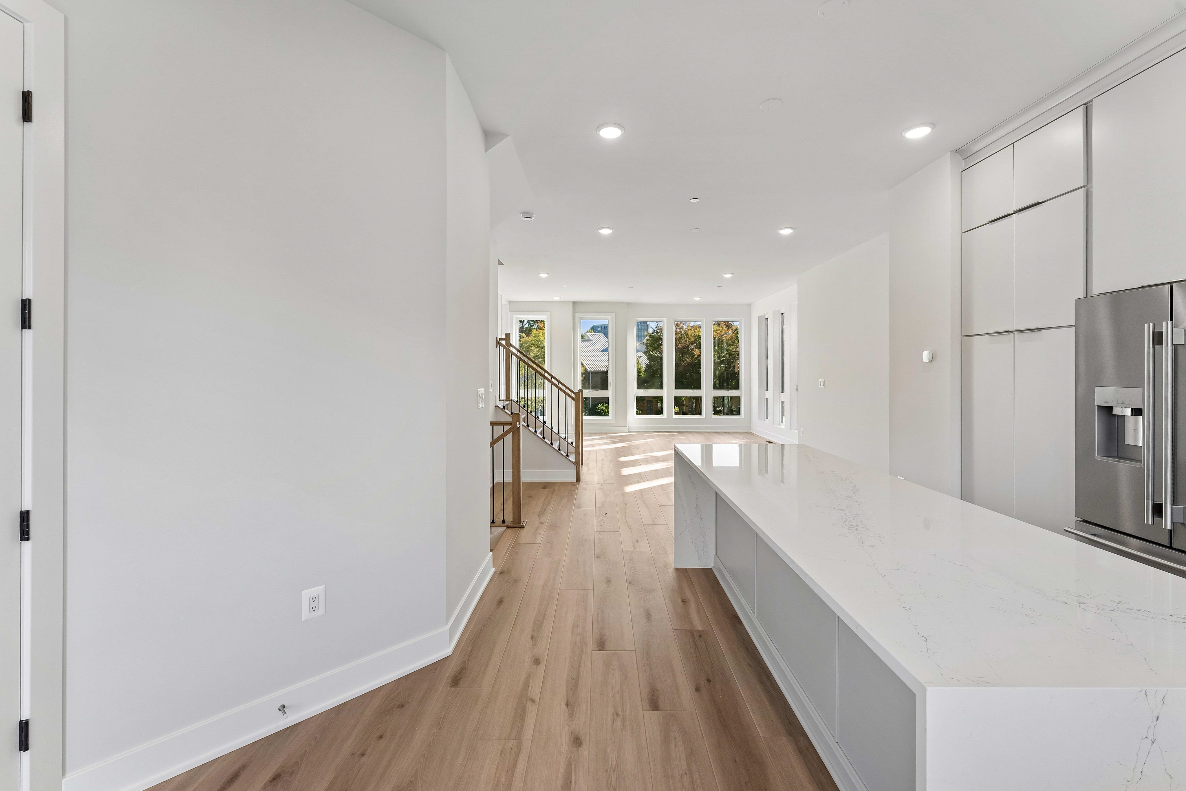 A modern white kitchen with a long marble island sits in the foreground, leading toward a bright living area and a staircase in the background.