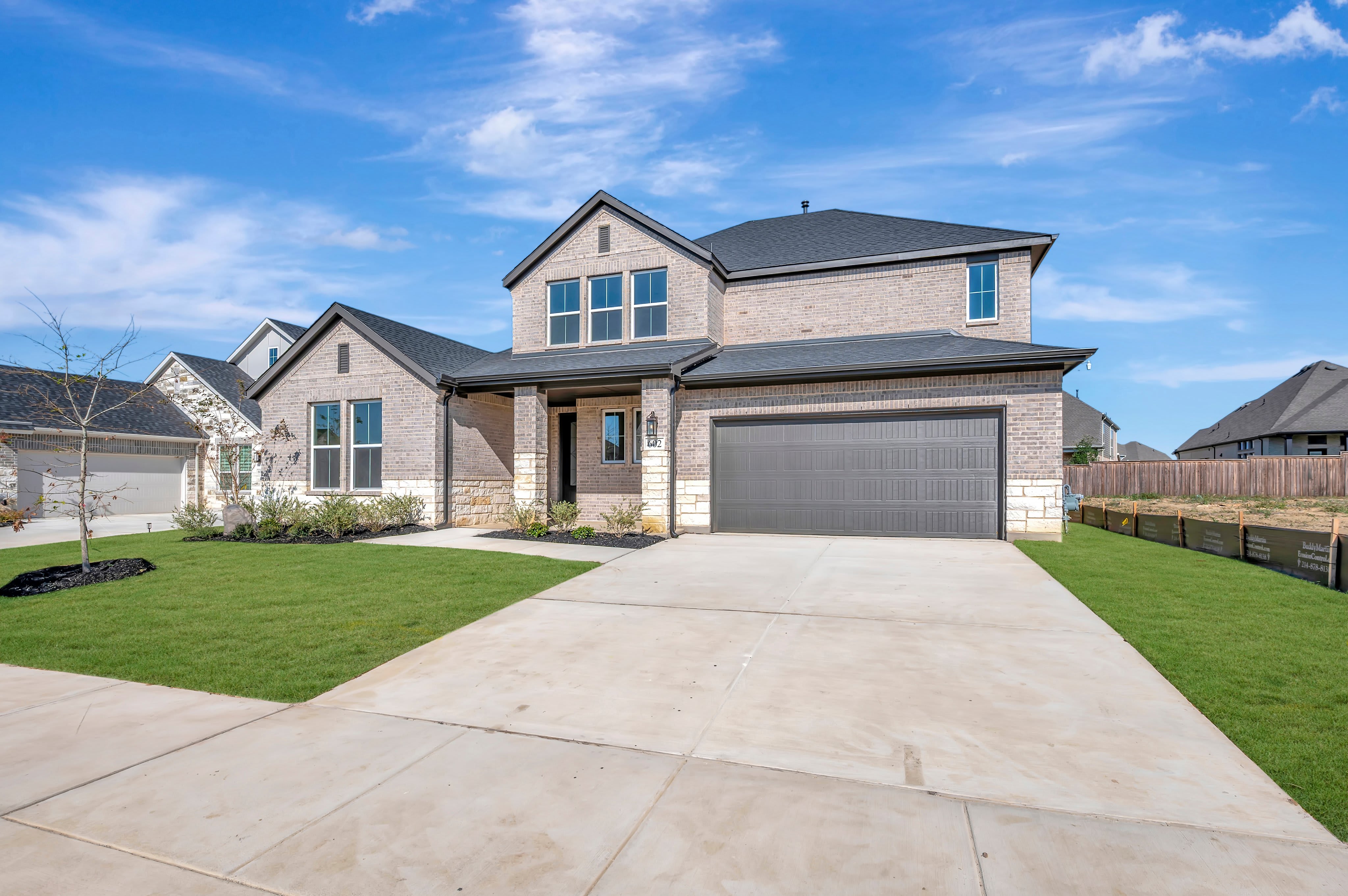 A modern two-story brick home with a grey garage door sits behind a concrete driveway and green lawn under a bright blue sky.