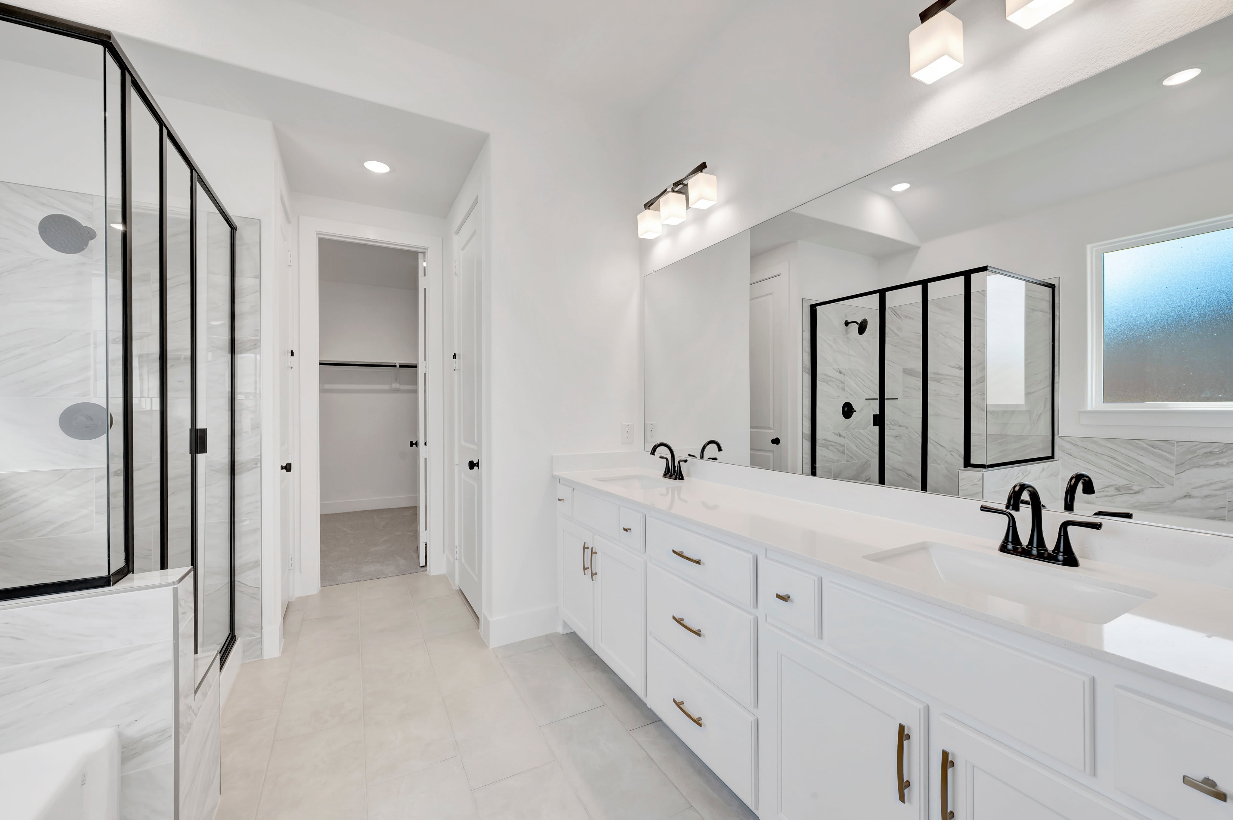 A modern white bathroom features a double vanity with black faucets in the foreground and a walk-in shower and open closet in the background.