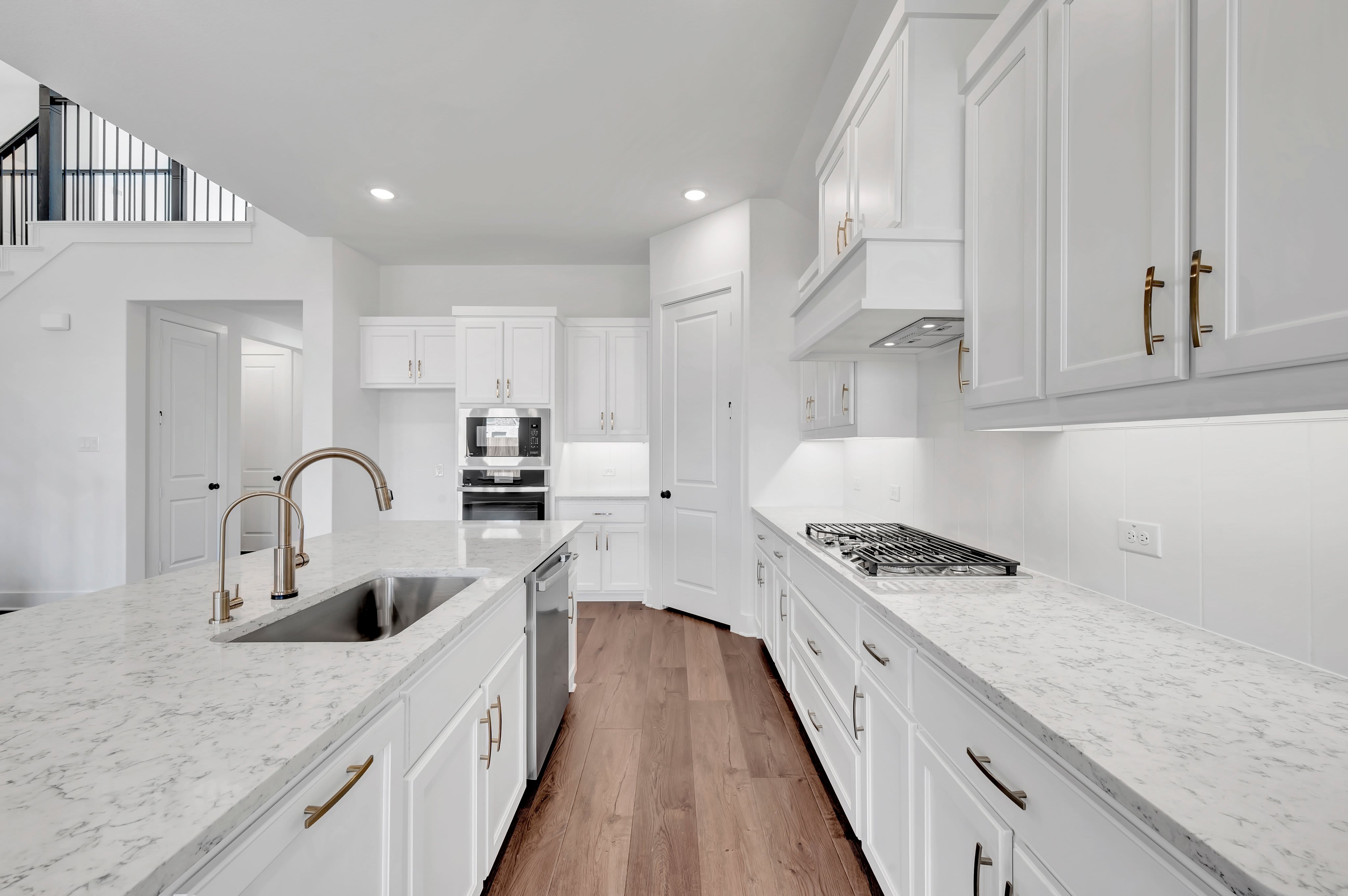 A modern white kitchen features a large marble island with a gold faucet in the foreground and white cabinetry with a gas stovetop in the background.