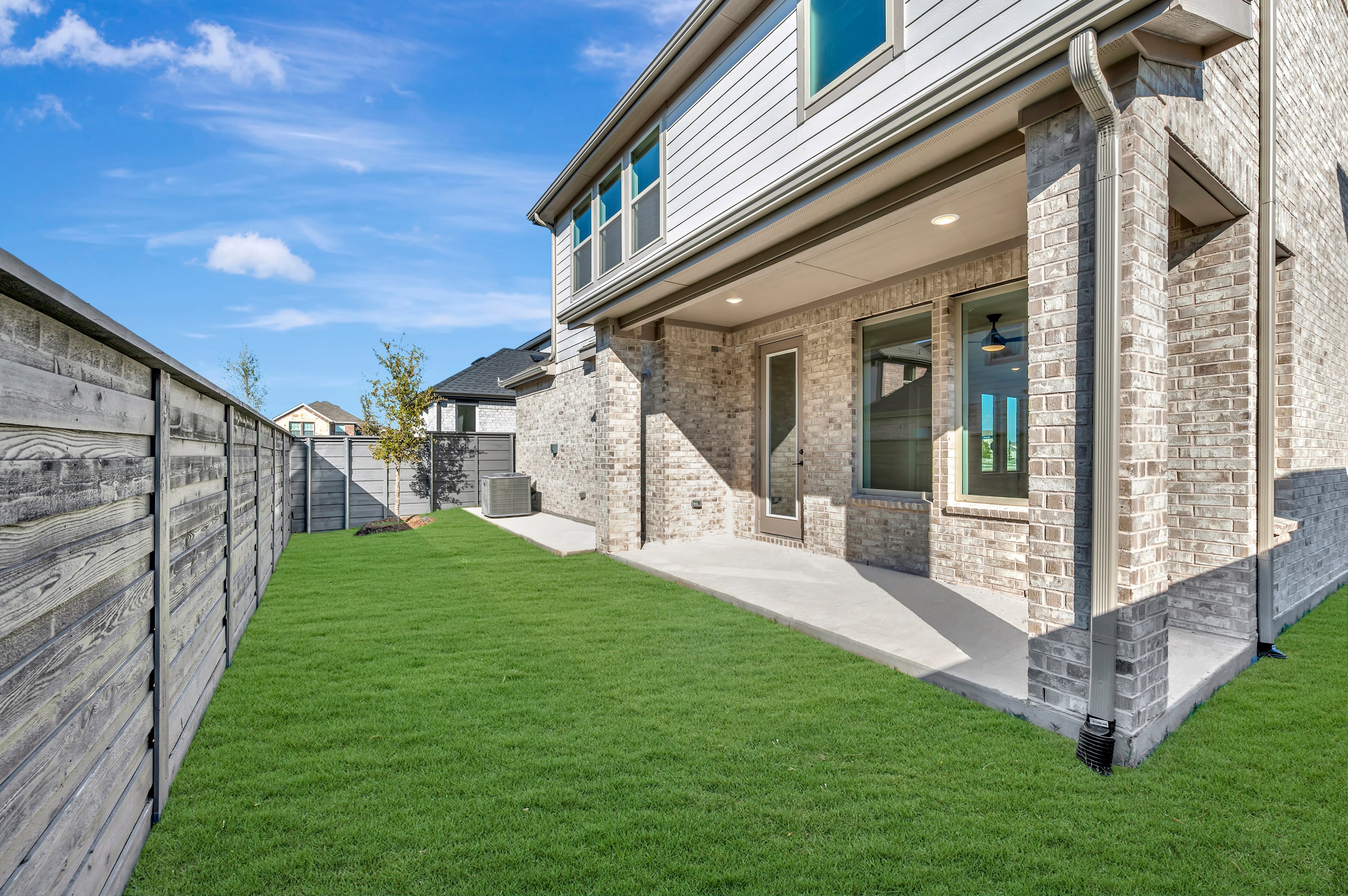 A green grassy lawn and wooden fence lead toward a concrete patio and the brick exterior of a modern house under a blue sky.