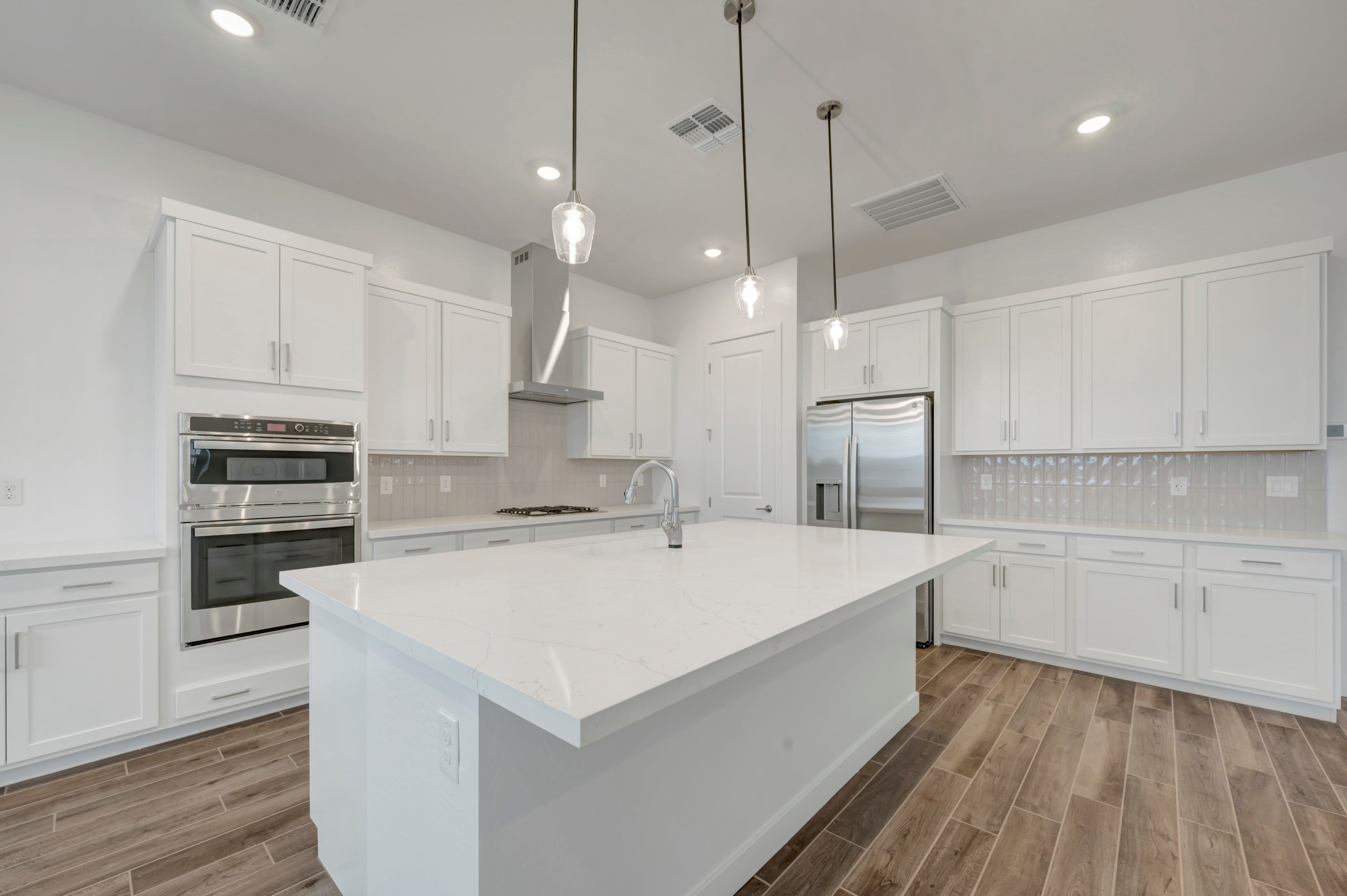 A large white kitchen island sits in the foreground of a modern kitchen featuring white cabinetry, stainless steel appliances, and wood-look flooring.