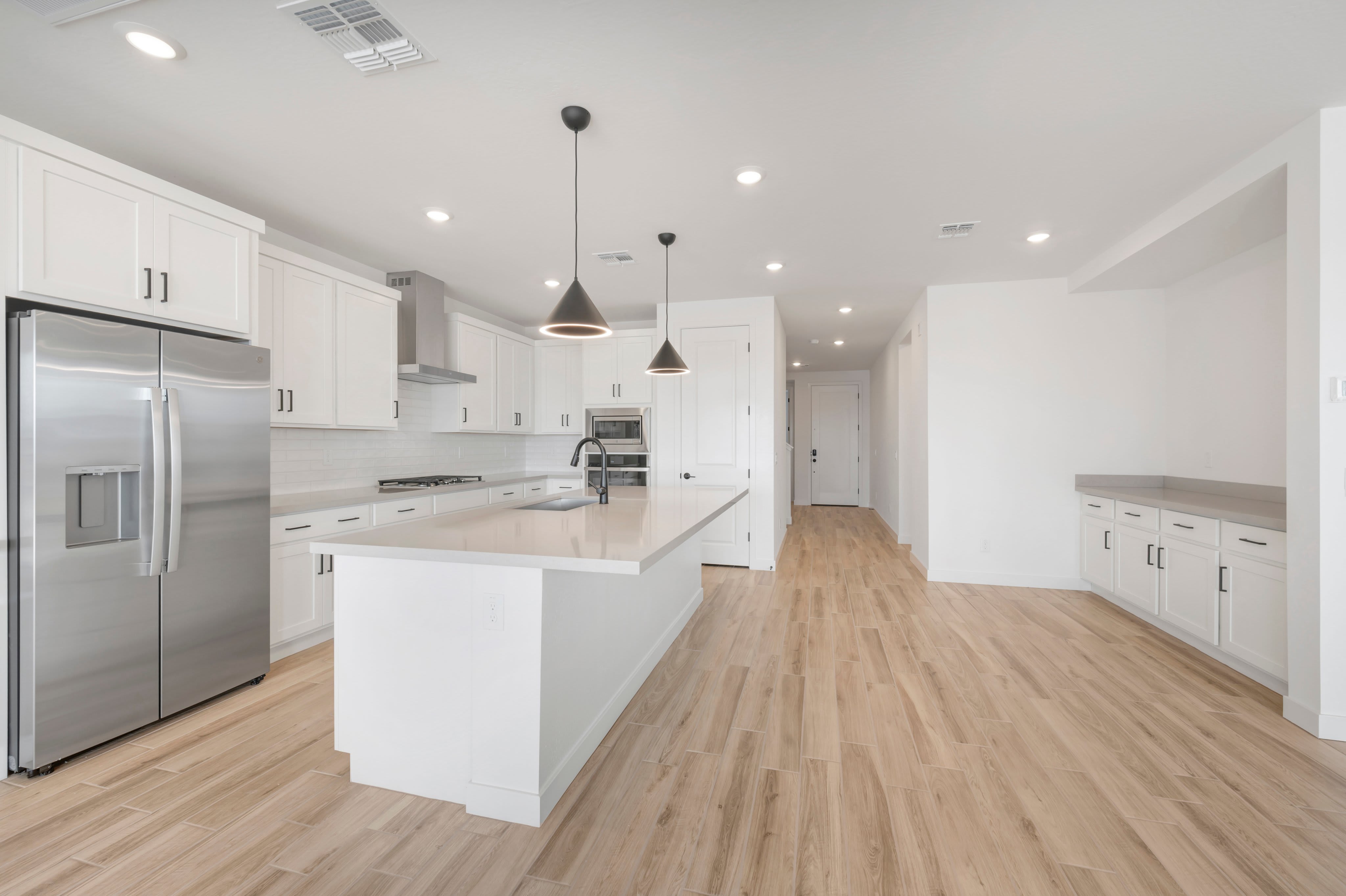 A modern white kitchen with a large central island and stainless steel appliances sits atop light wood flooring in an open-concept living space.