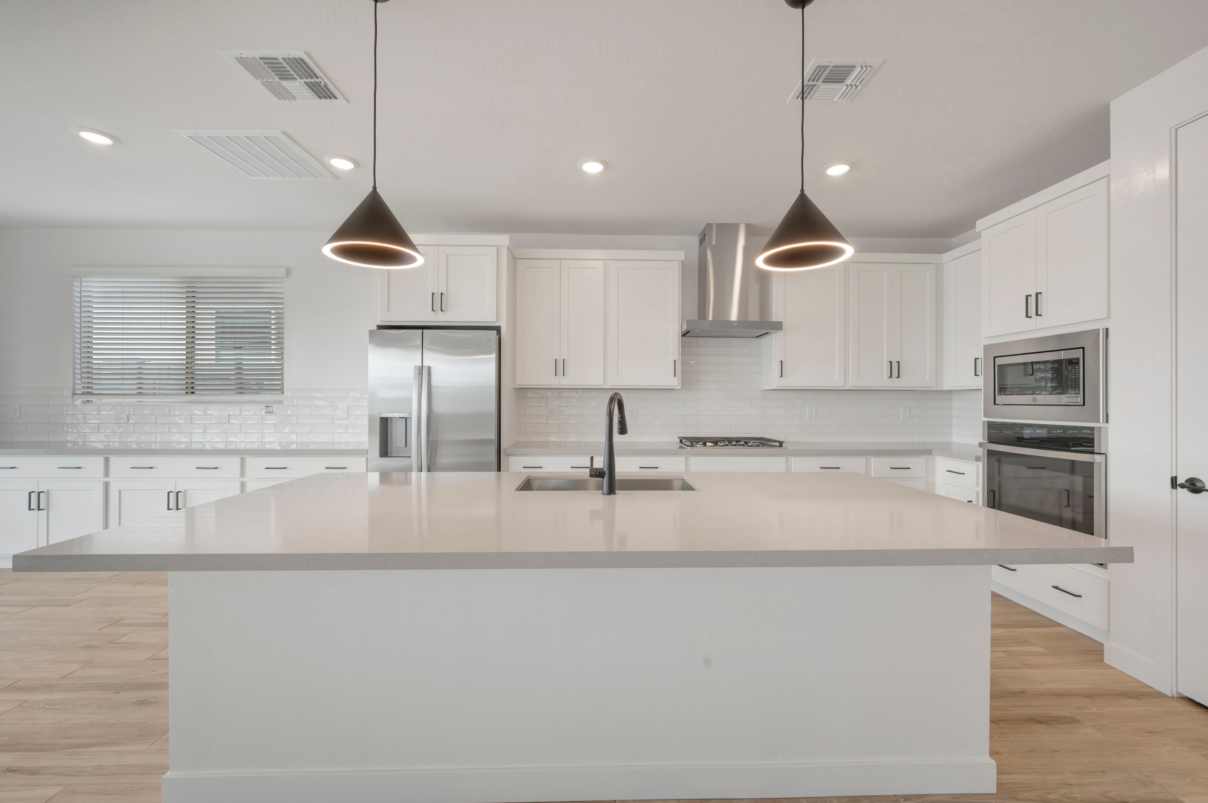 A large white kitchen island sits in the foreground of a modern, all-white kitchen featuring stainless steel appliances and cone-shaped pendant lights.