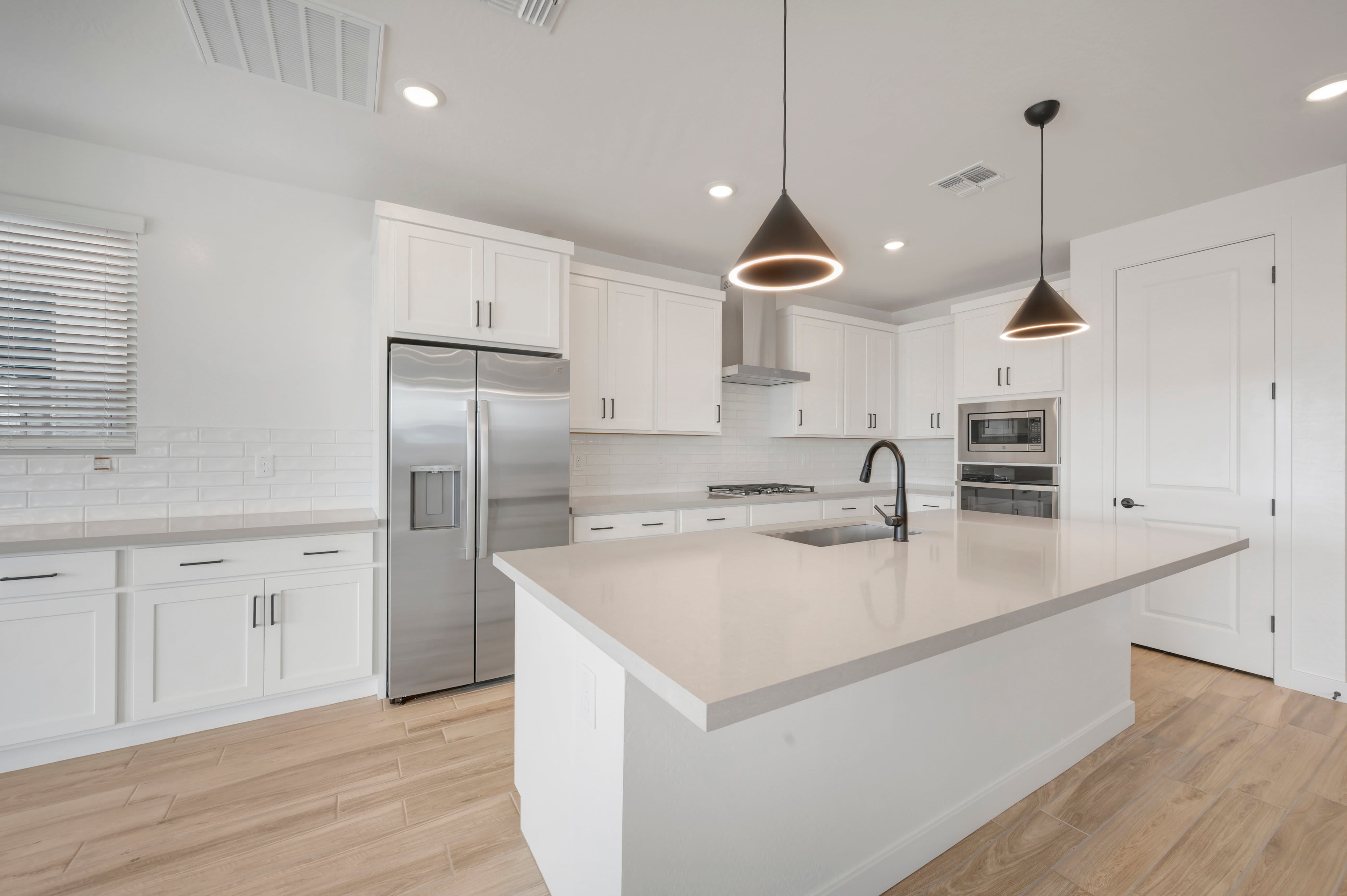A white kitchen island with a sink and black faucet sits in the foreground of a modern room featuring white cabinetry, stainless steel appliances, and light wood flooring.