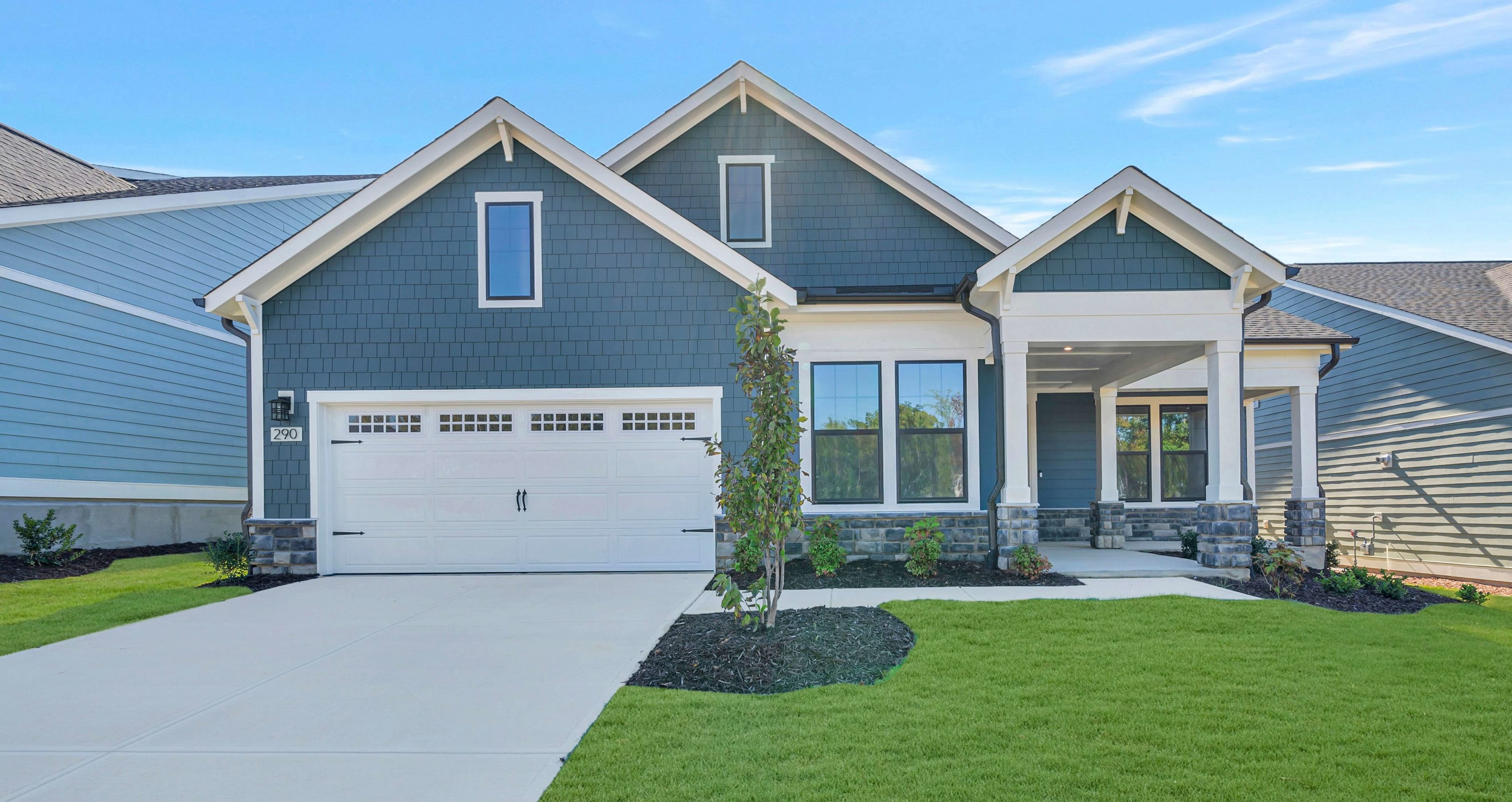 A blue house with a white garage door and a concrete driveway sits behind a manicured green lawn under a clear blue sky.