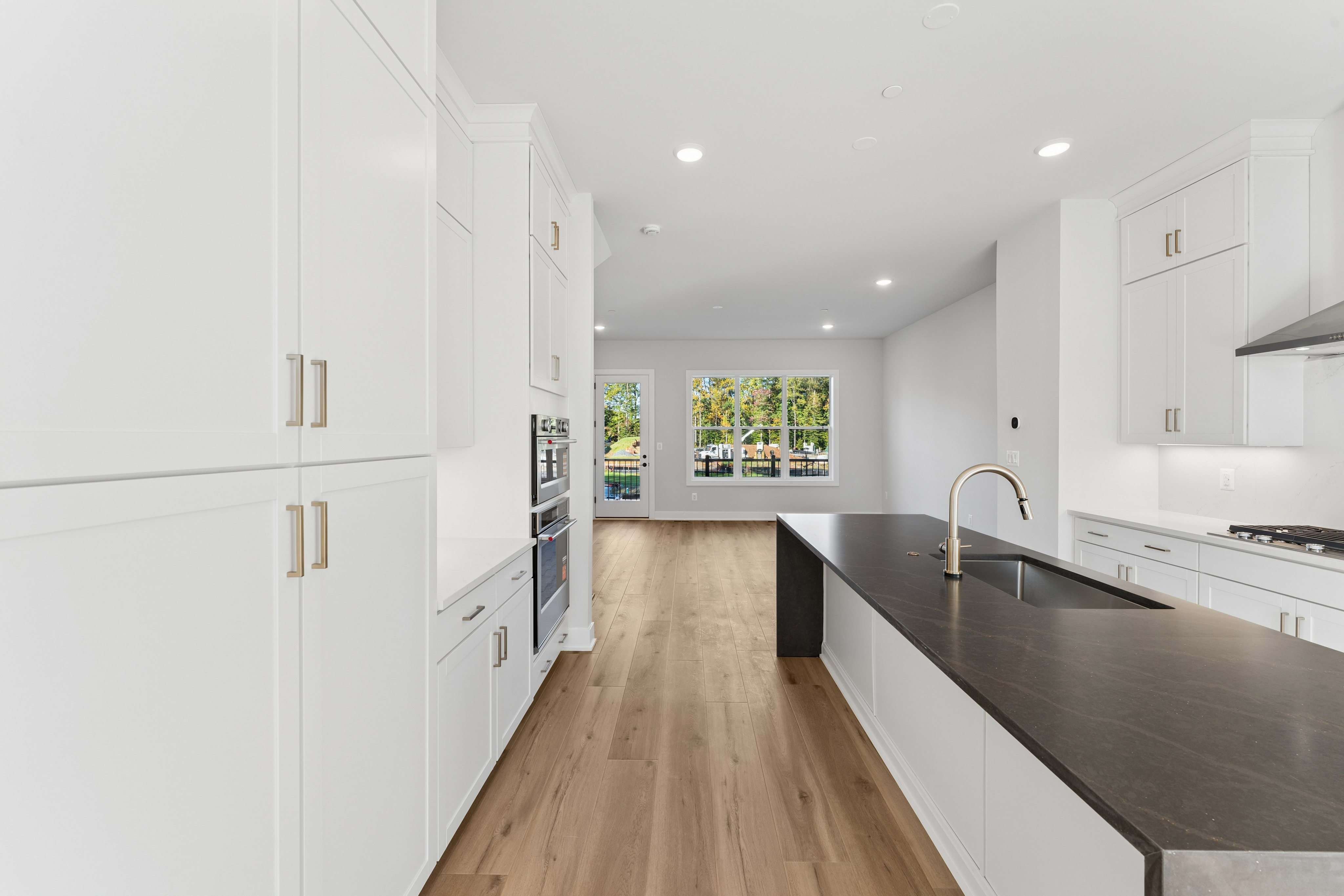 White kitchen cabinets and a dark island with a sink occupy the foreground, leading toward a bright living area with a glass door and window in the background.