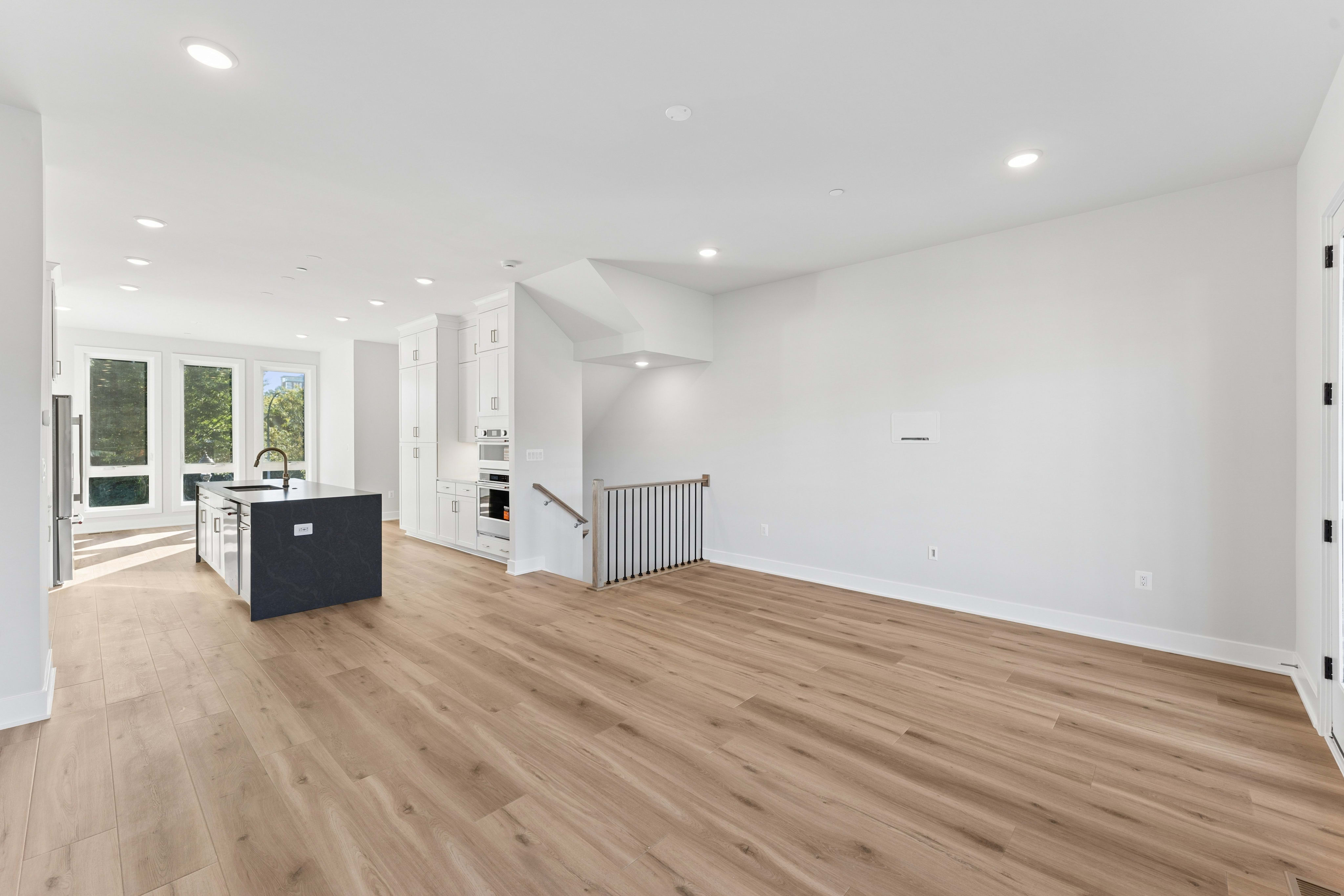 An open-concept living area with light wood flooring in the foreground leads to a modern kitchen with white cabinetry and a dark island in the background.