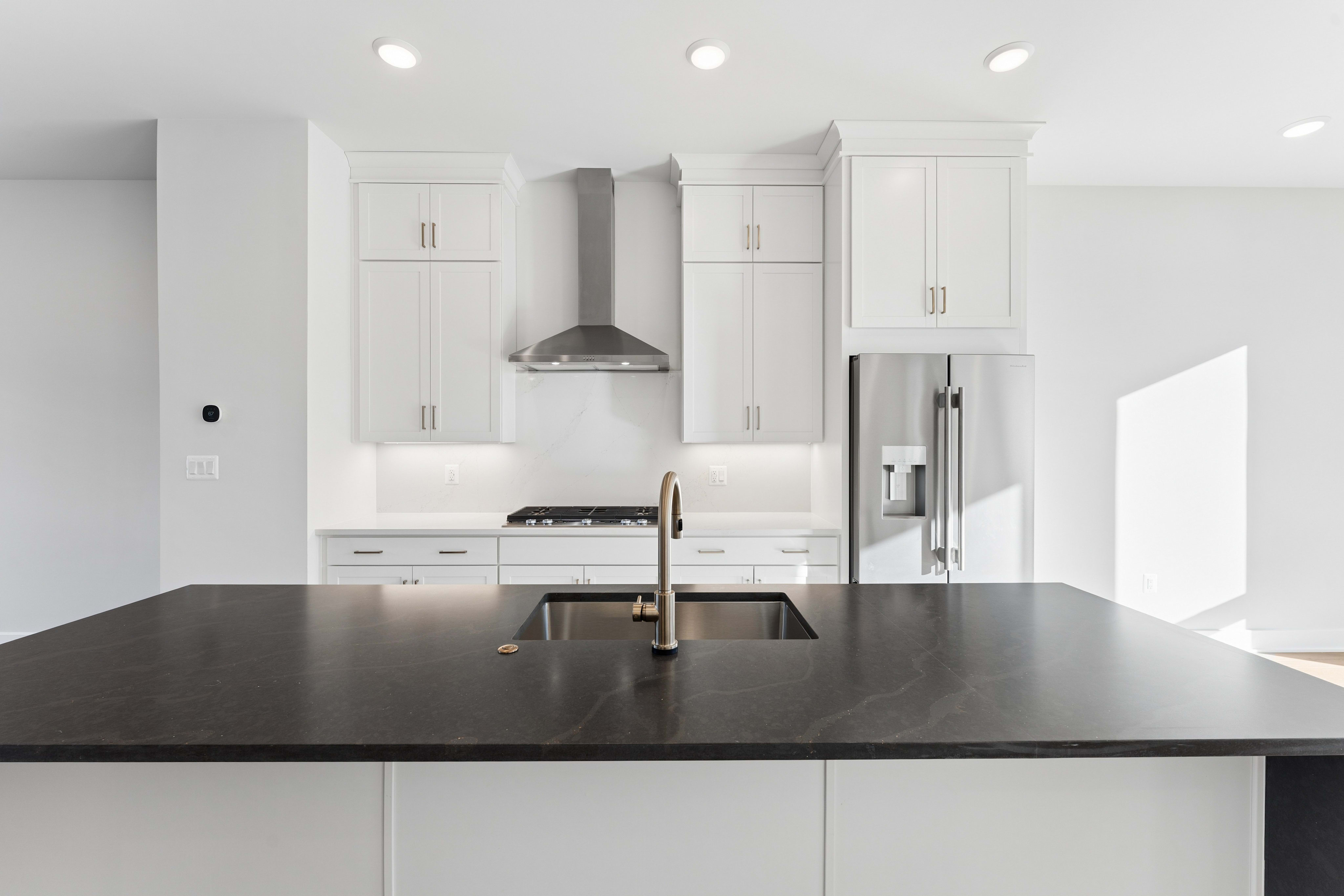A dark grey kitchen island with a sink and faucet sits in the foreground of a bright white kitchen featuring white cabinetry and stainless steel appliances.
