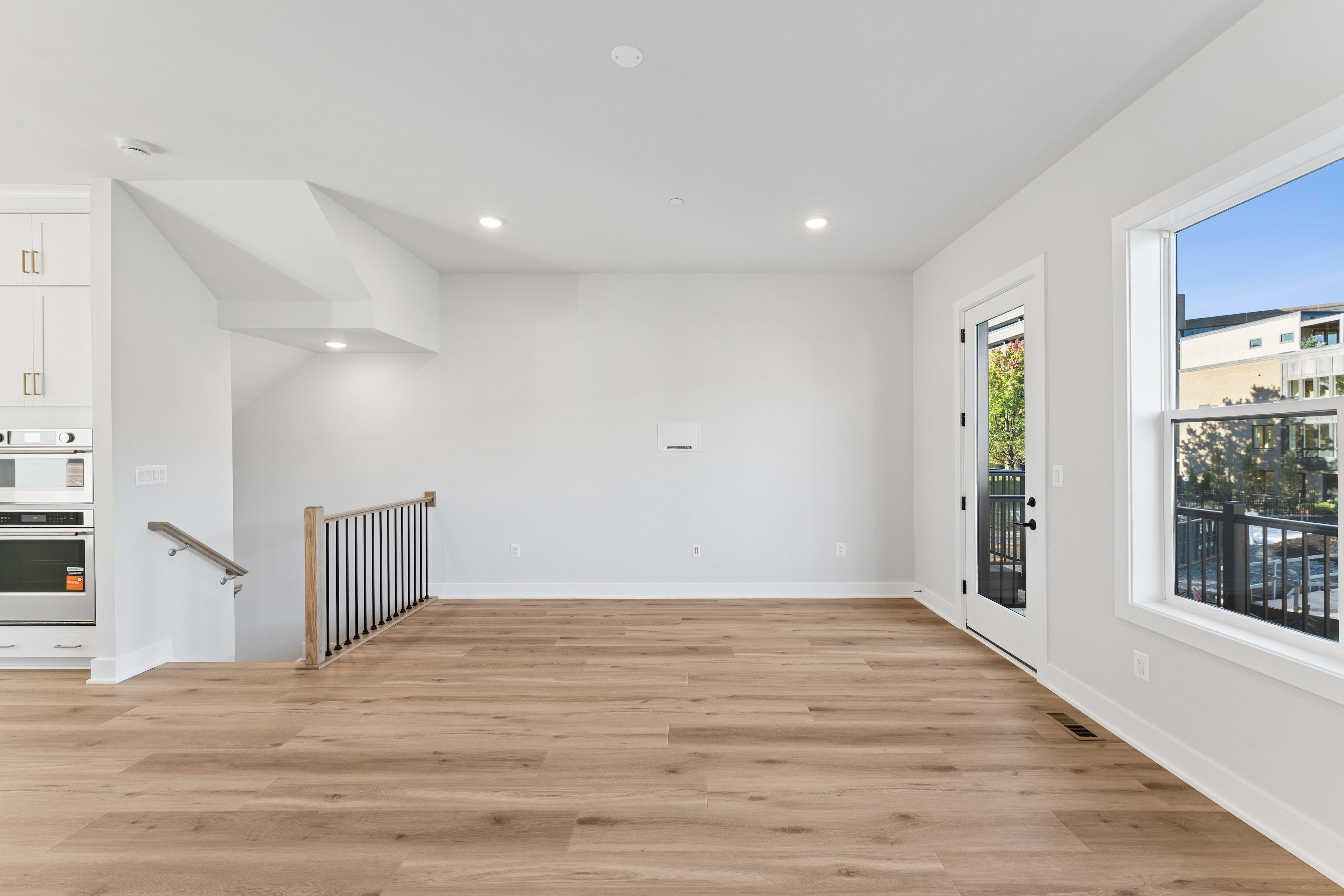 Light-colored hardwood floors fill the foreground of an empty white room featuring a staircase on the left and a glass door and window on the right.