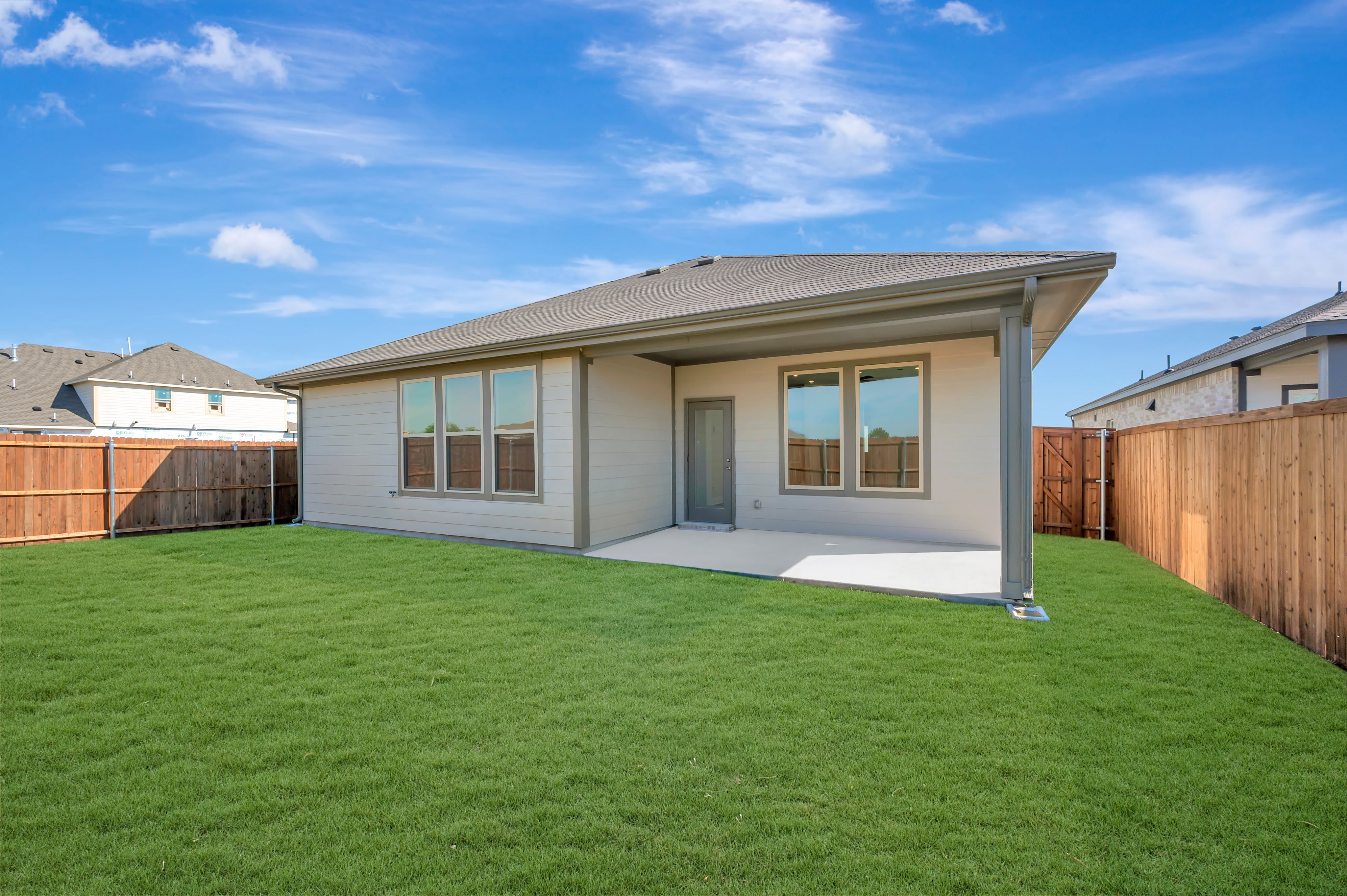 A lush green lawn leads to a modern light-colored house with a concrete patio, set against a bright blue sky with scattered clouds.