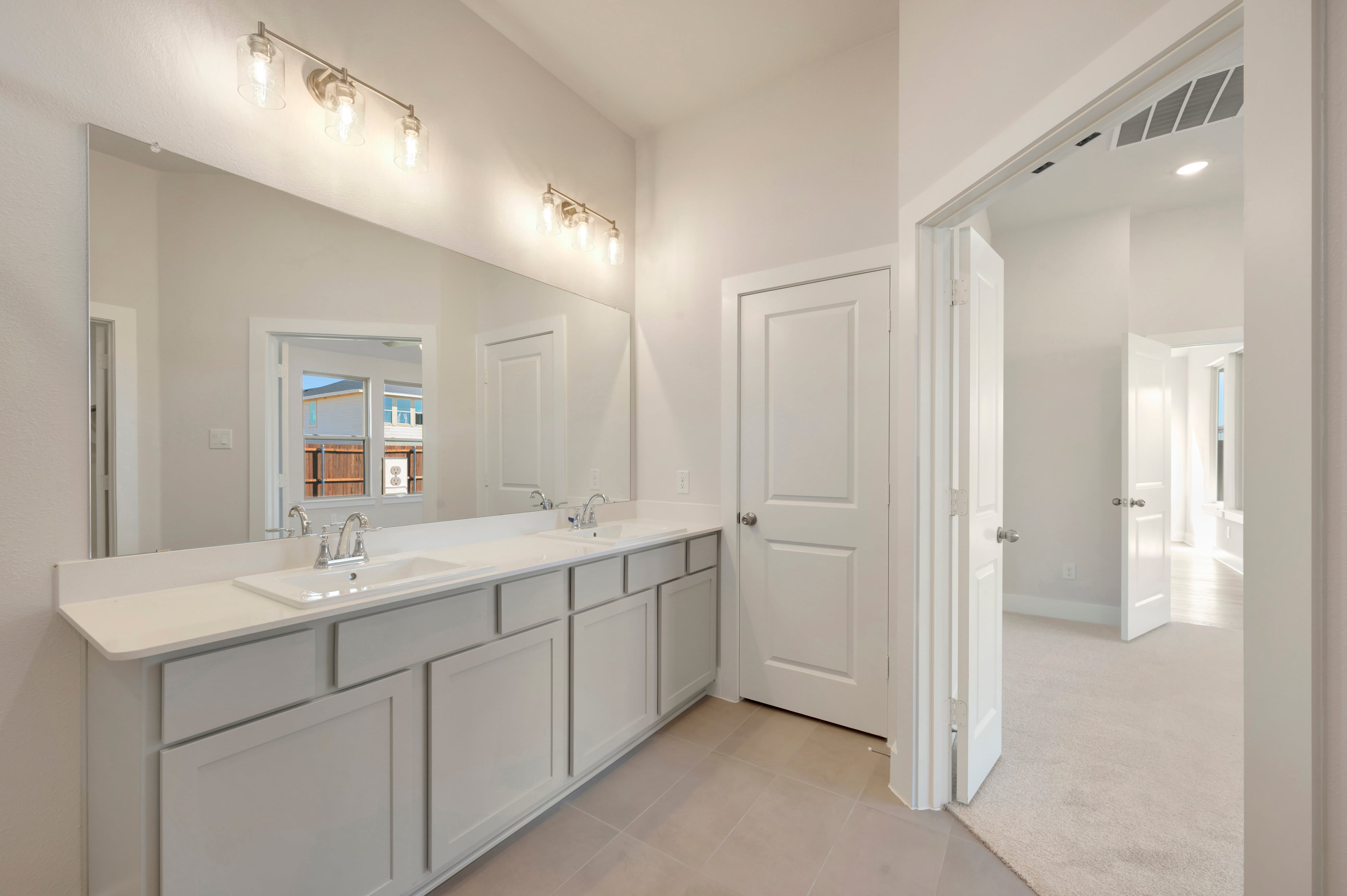 A white double vanity with a large mirror sits in the foreground of a bright bathroom, with an open doorway leading to a carpeted hallway in the background.