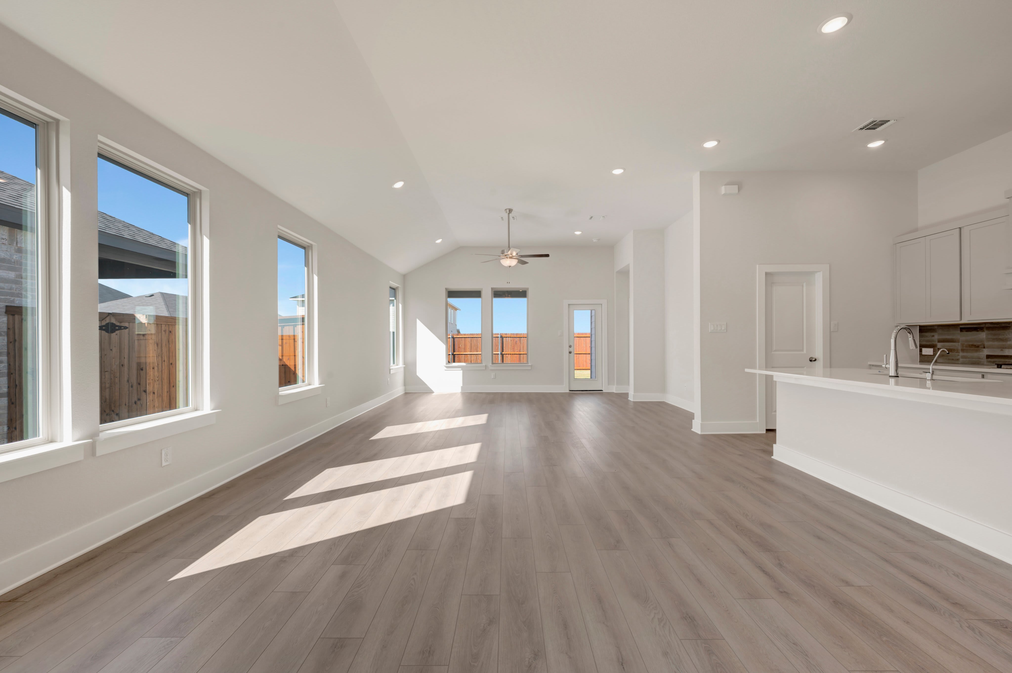 An empty open-concept living area with light wood flooring in the foreground leads to a bright kitchen and a sunlit room with a ceiling fan in the background.