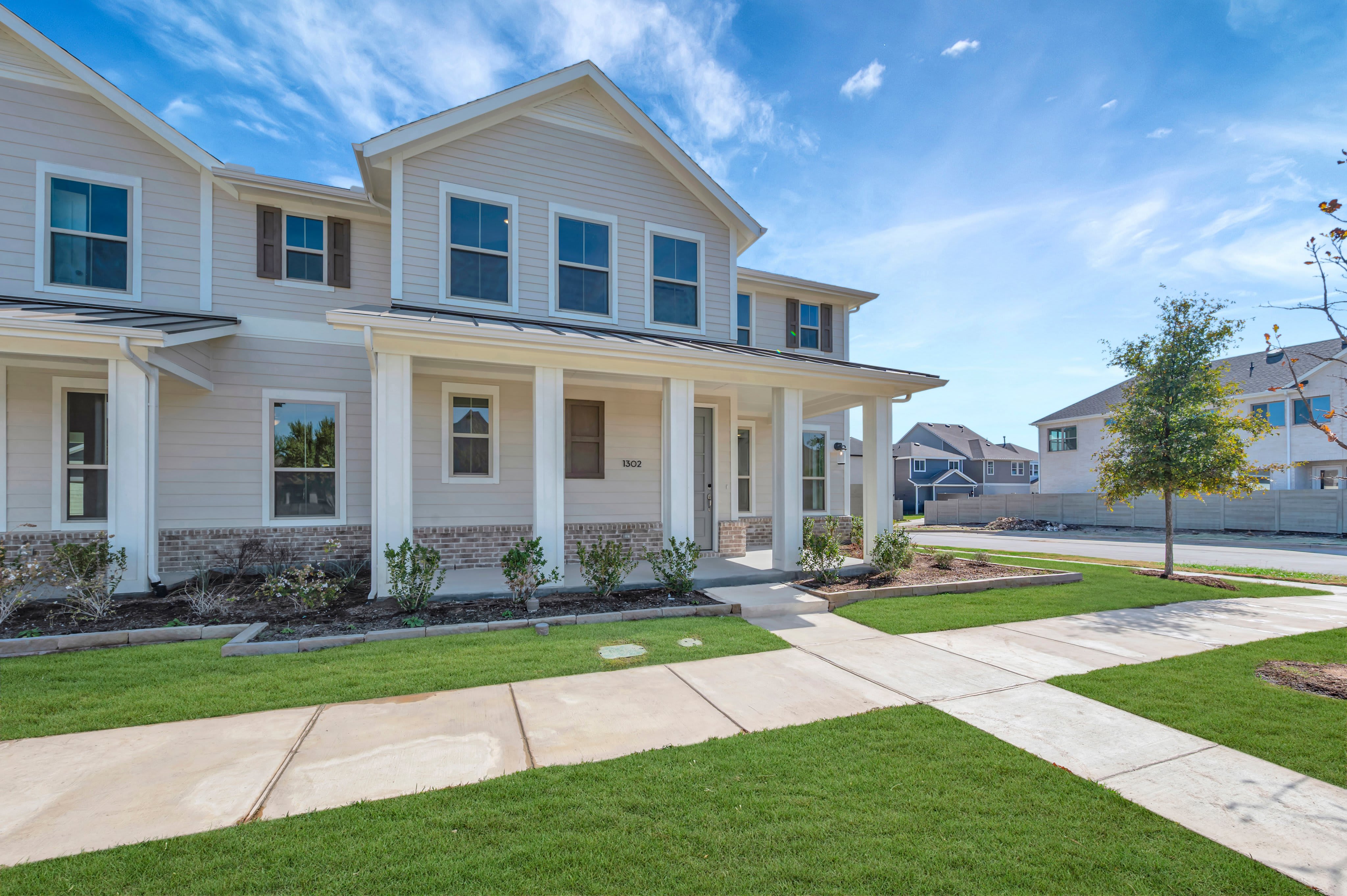A two-story beige house with a white porch sits behind a green lawn and concrete walkway under a bright blue sky.