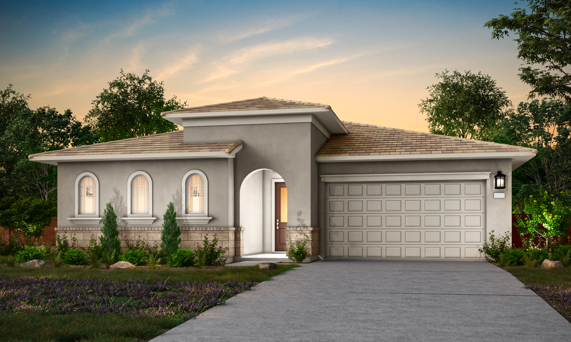 A single-story, Mediterranean-style house with a tiled roof, surrounded by lush greenery and a paved driveway leading to the front entrance, set against a picturesque sunset sky.