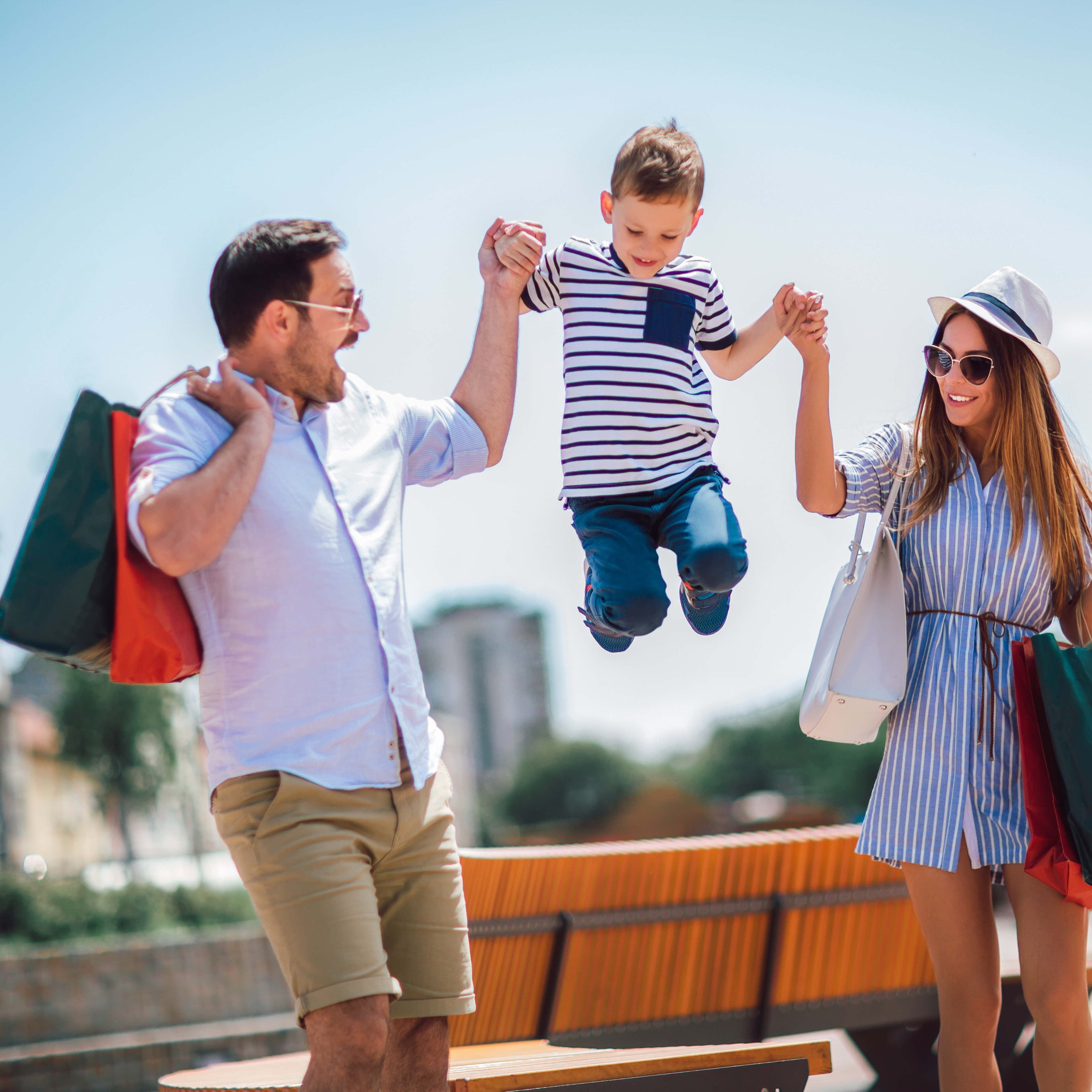 A group of young people, a man and two women, are walking together and one of the women is jumping up in the air, holding shopping bags, against a backdrop of buildings and a sunny outdoor setting.
