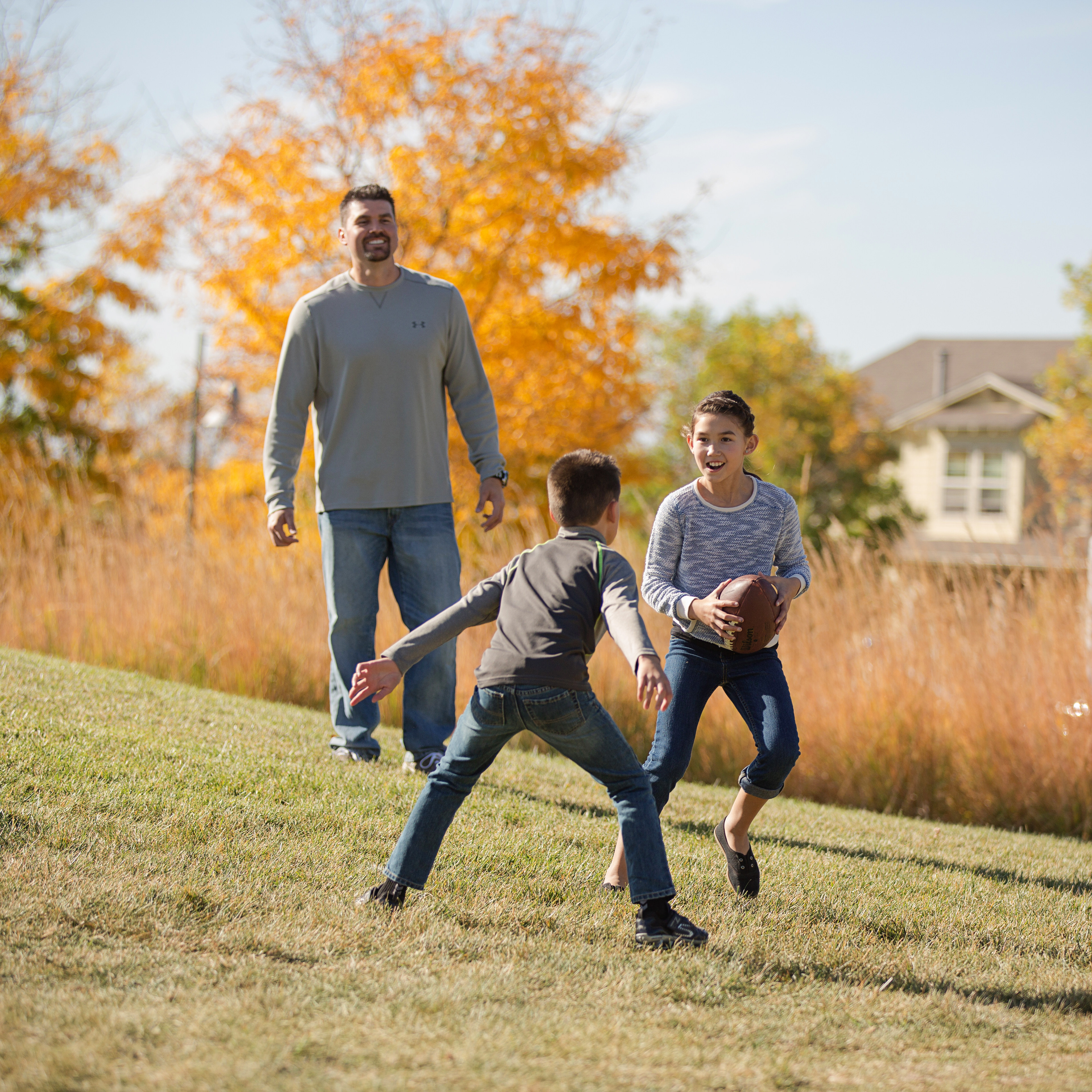 A family of three walking together on a grassy path surrounded by vibrant autumn foliage and a residential neighborhood in the background.