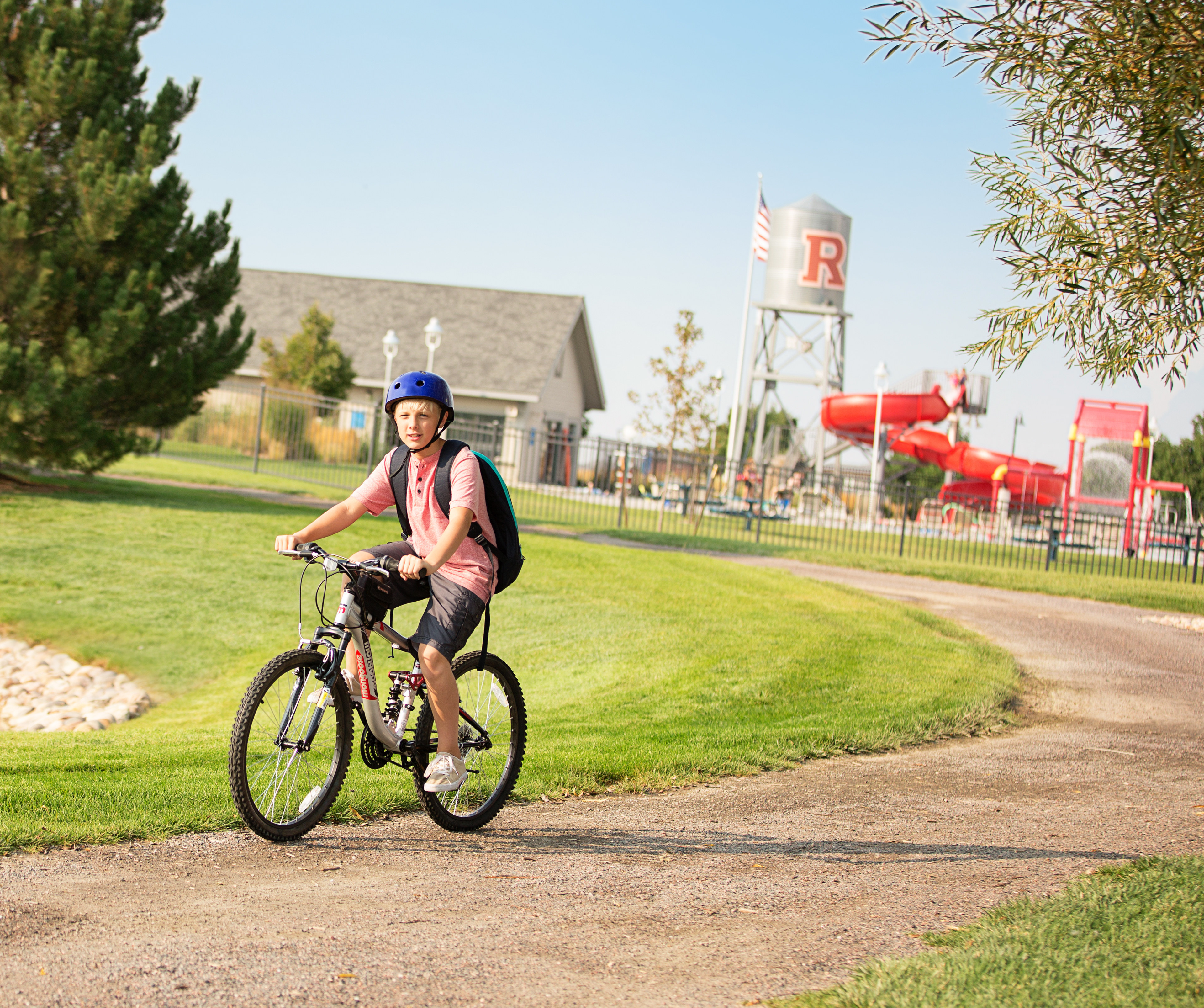 A person wearing a blue hat and a pink jacket is riding a bicycle on a dirt path surrounded by lush greenery, with buildings and signage visible in the background.