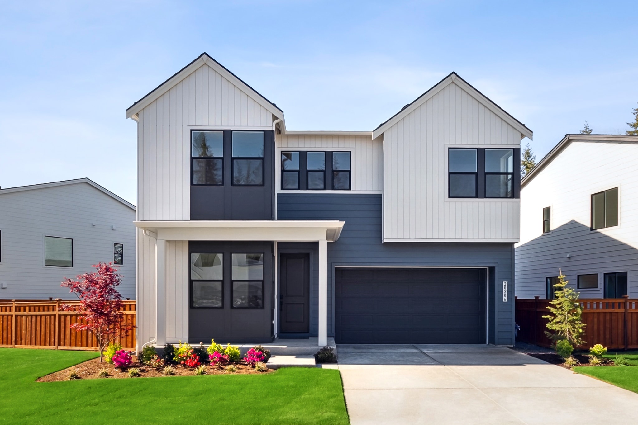 A modern two-story house with a white exterior, black trim, and a well-manicured lawn with colorful flowers in the foreground, set against a clear blue sky.
