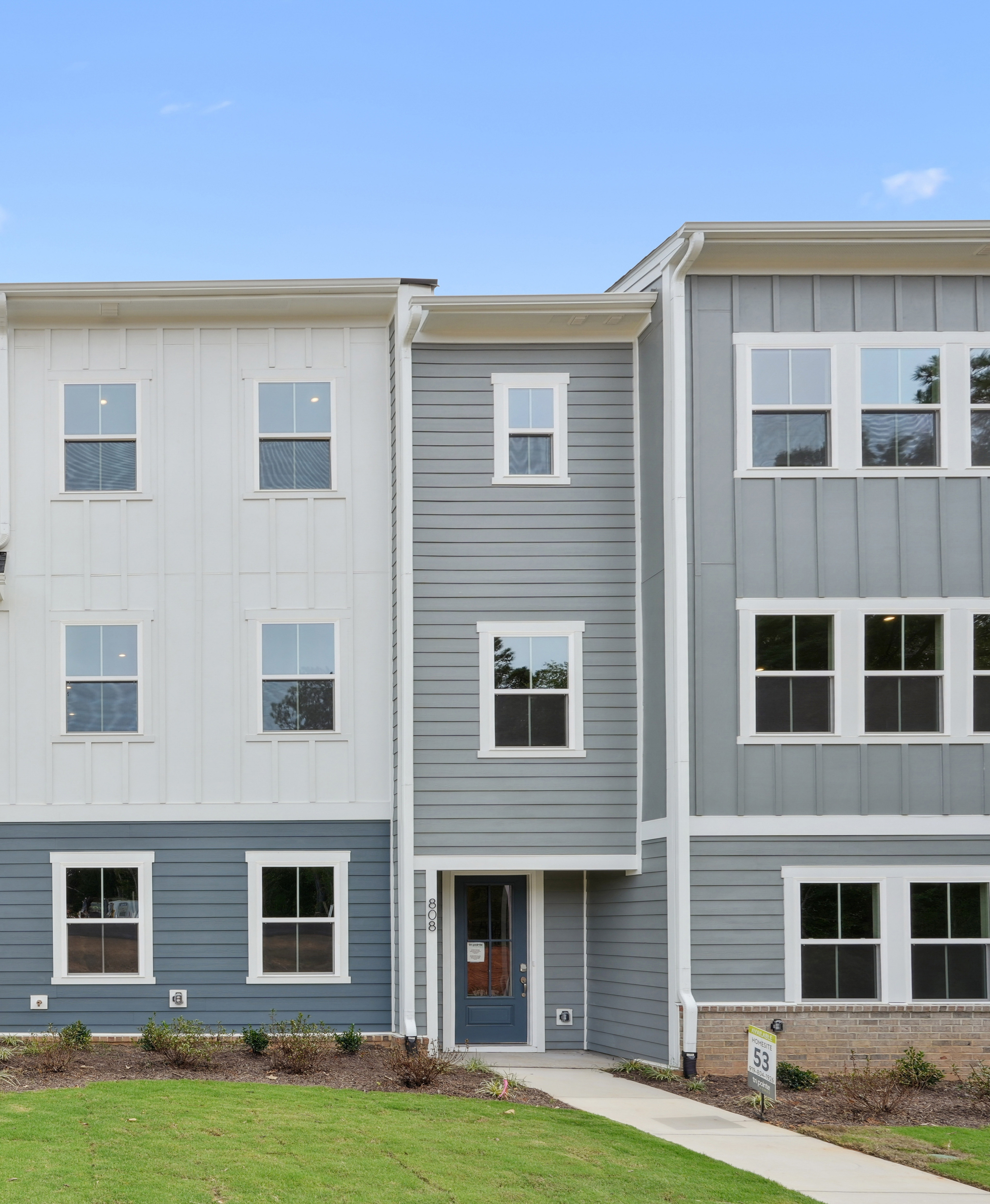 A multi-story townhouse complex with gray siding, white trim, and a grassy lawn in the foreground.
