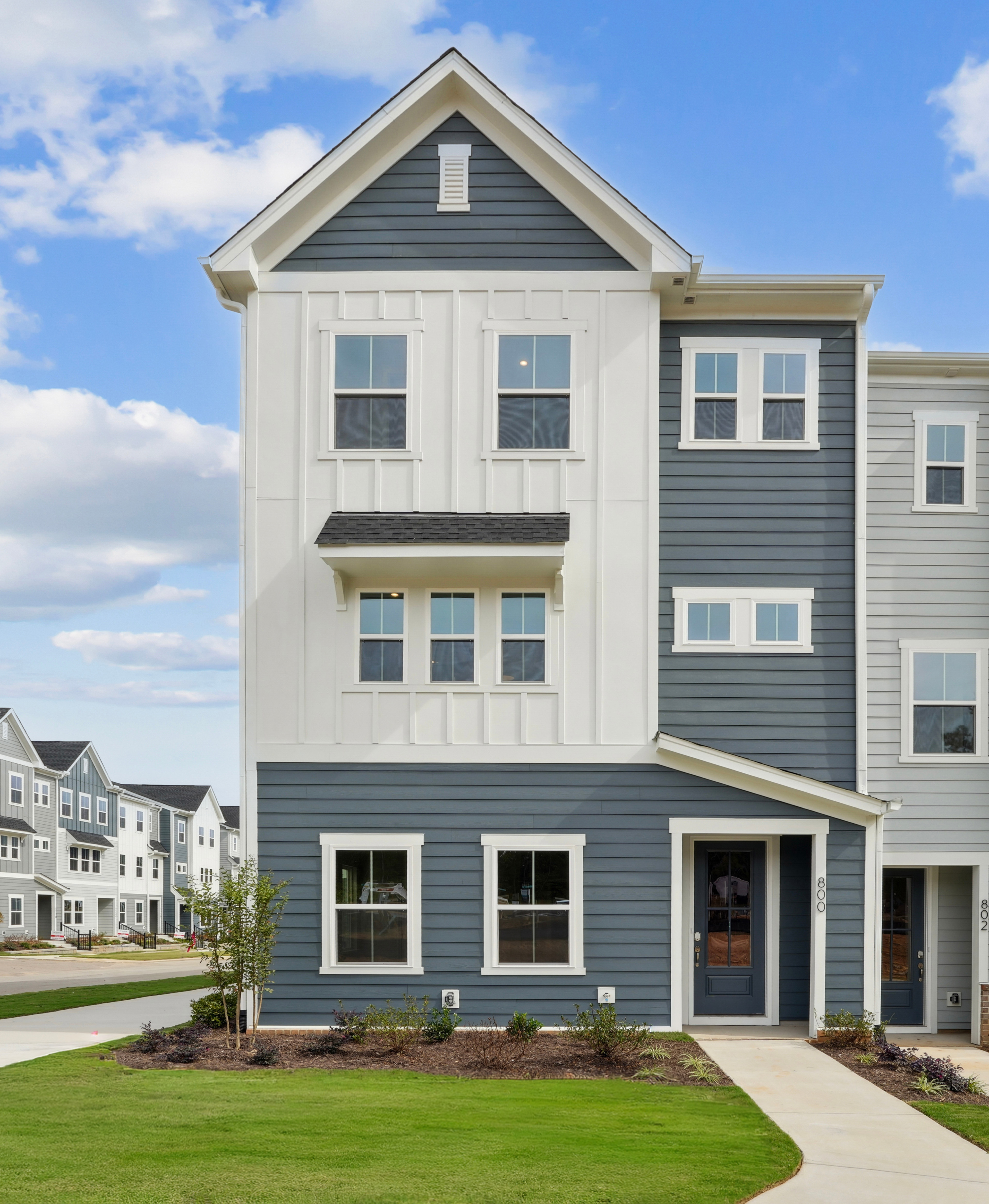 A multi-story residential building with a gray exterior and white trim, surrounded by a well-manicured lawn and set against a blue sky with scattered clouds.
