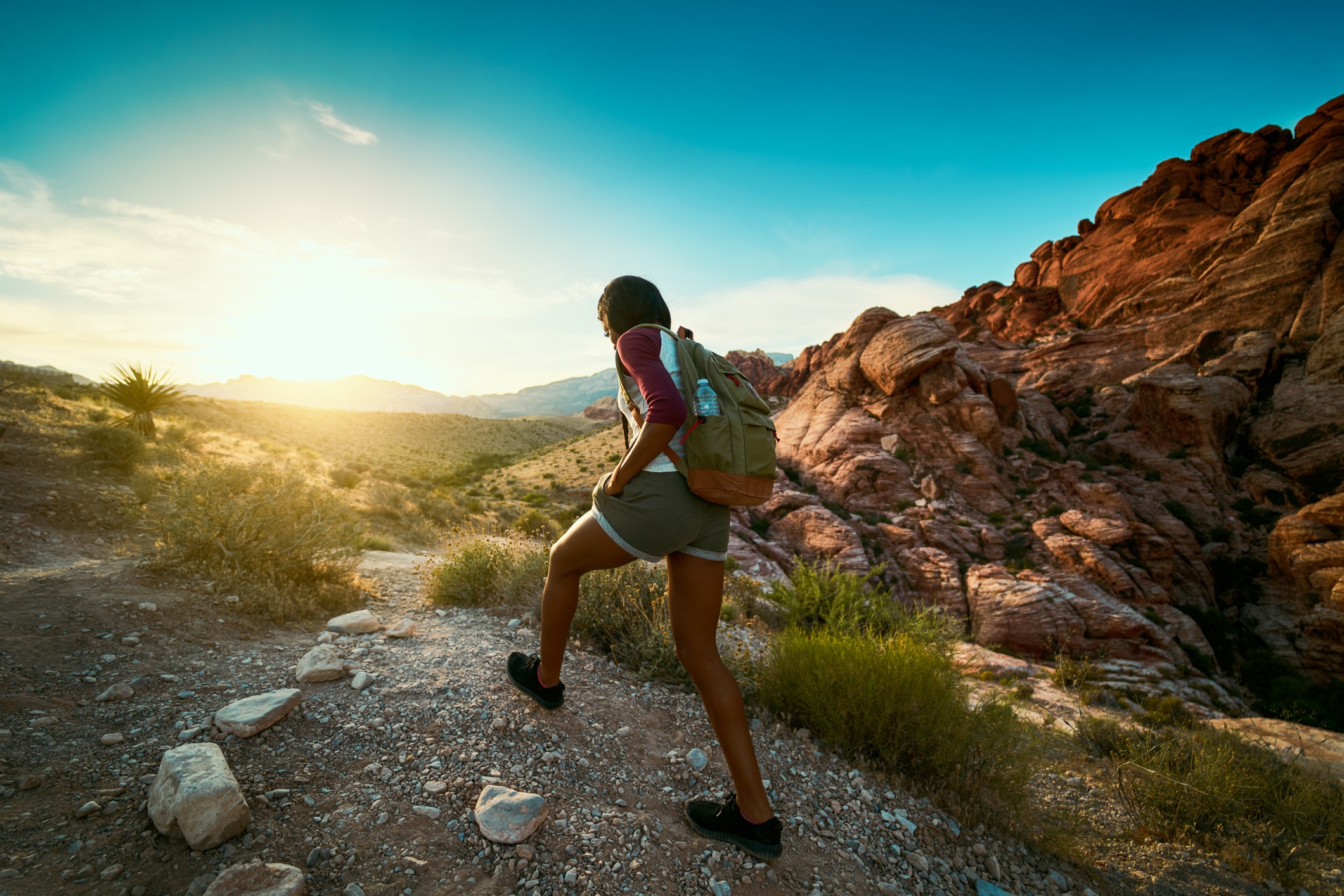 A person in hiking gear walks along a rocky trail in a scenic desert landscape, with mountains and a bright sky in the background.