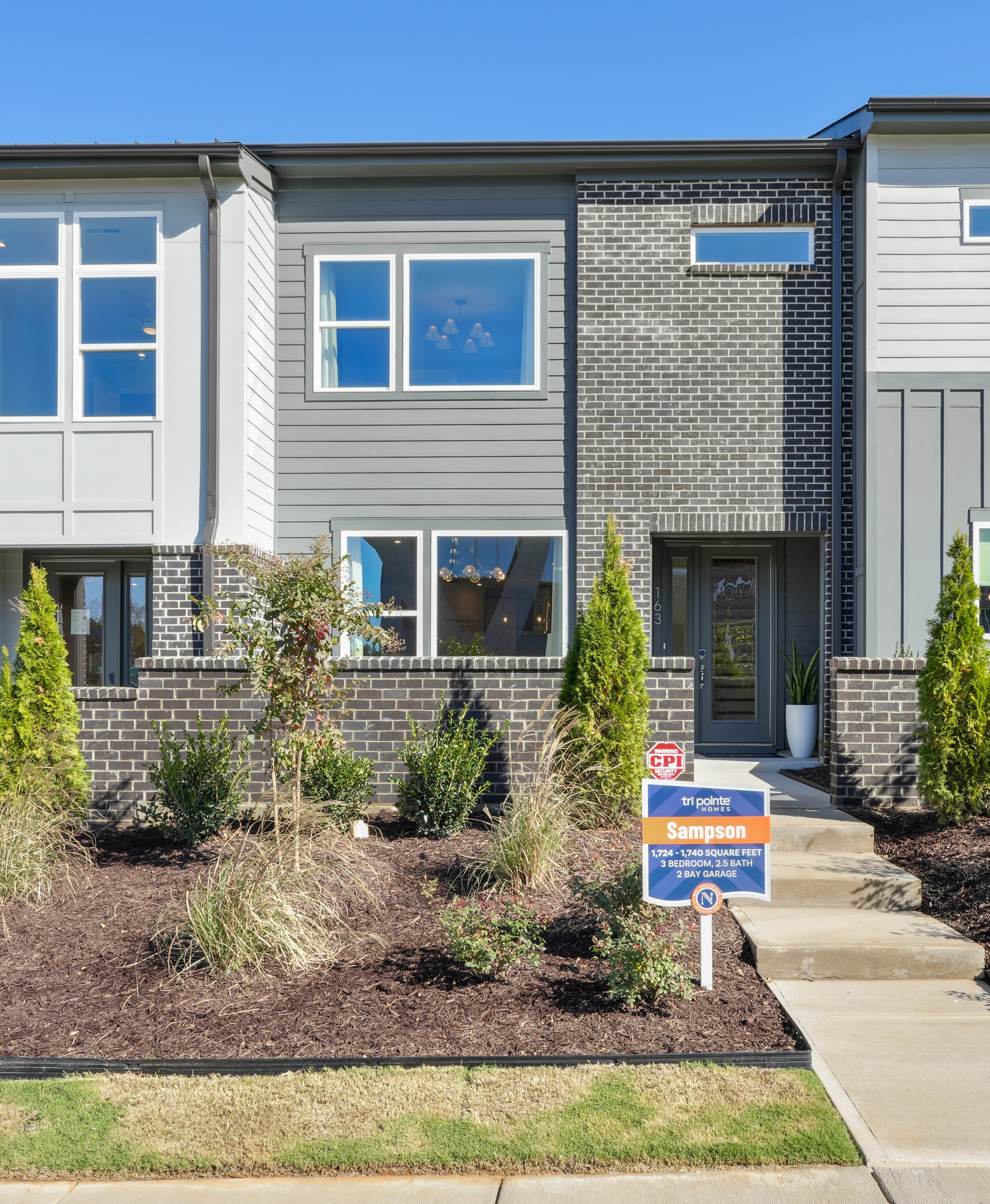A modern townhouse with a well-landscaped front yard, featuring a brick and siding exterior, large windows, and a paved walkway leading to the entrance.