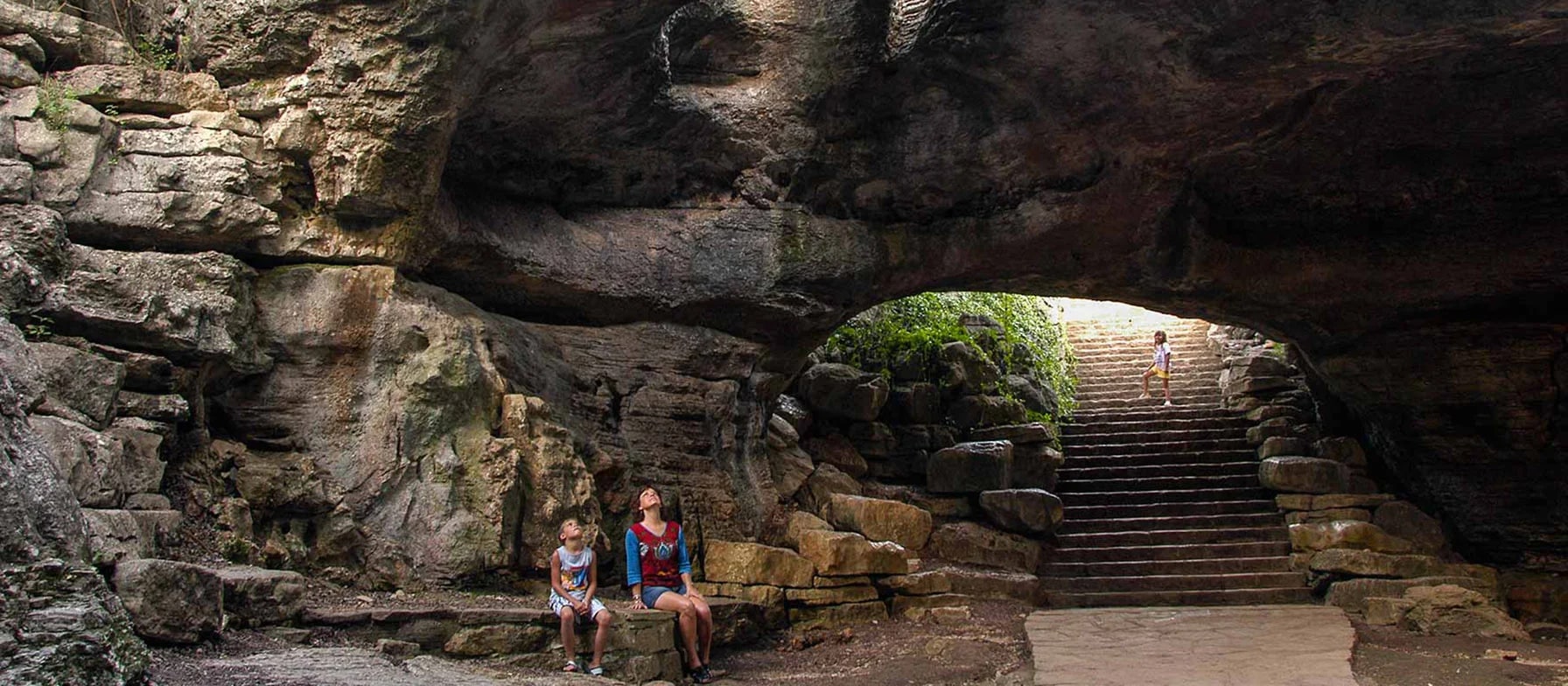 A rocky, cave-like structure with a stone archway leading to a staircase, with two people standing in the foreground.