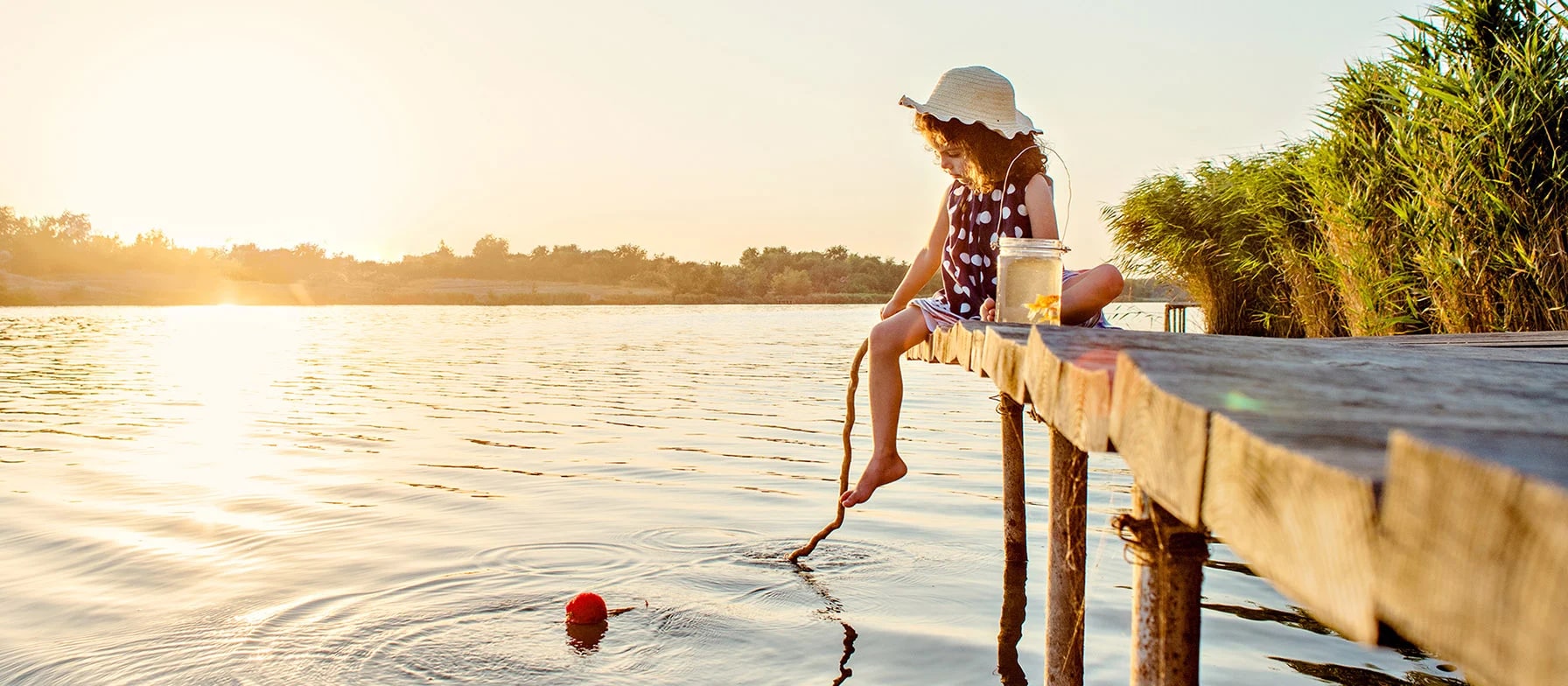 A person in a colorful dress stands on a wooden dock, gazing out over a serene lake surrounded by lush greenery as the sun sets in the distance.