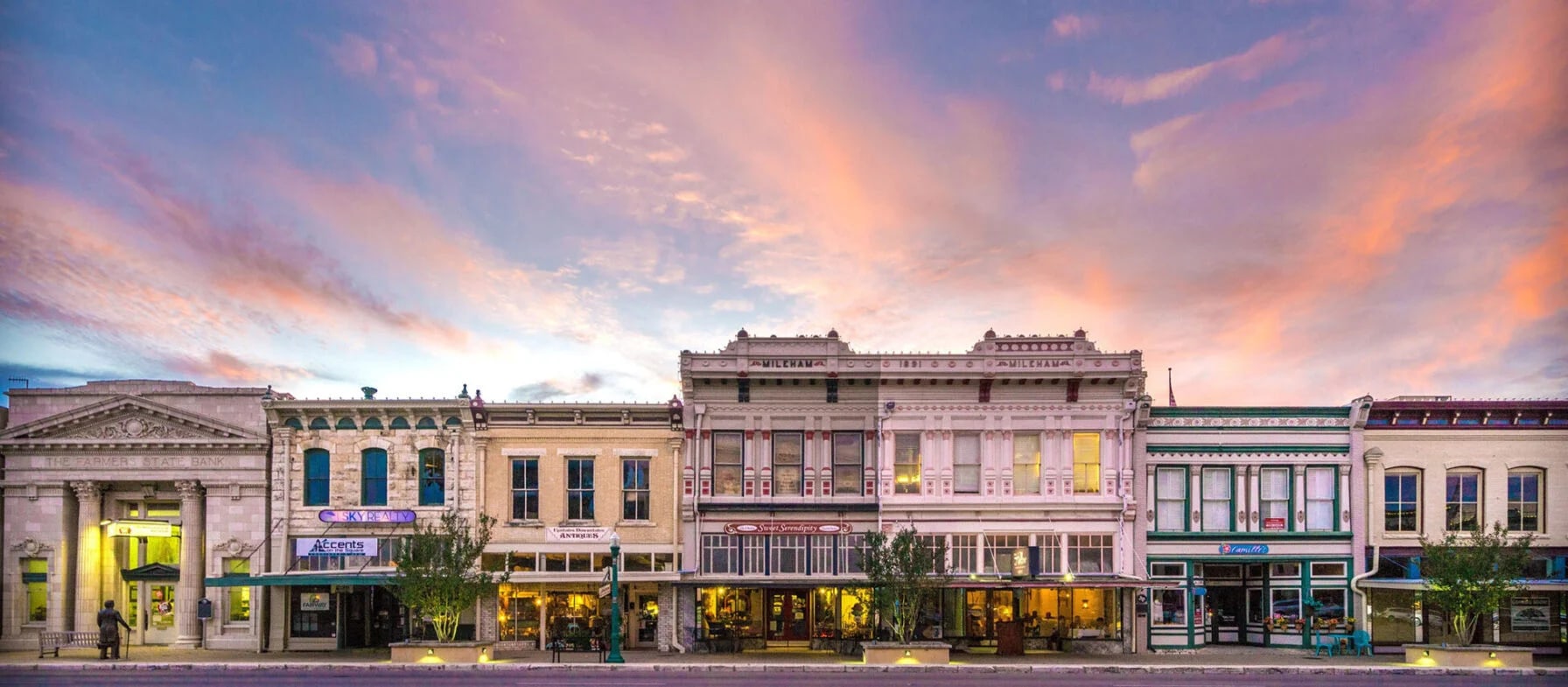 A vibrant and colorful cityscape with a row of historic buildings, shops, and restaurants against a dramatic sky filled with clouds in warm hues of orange and pink.