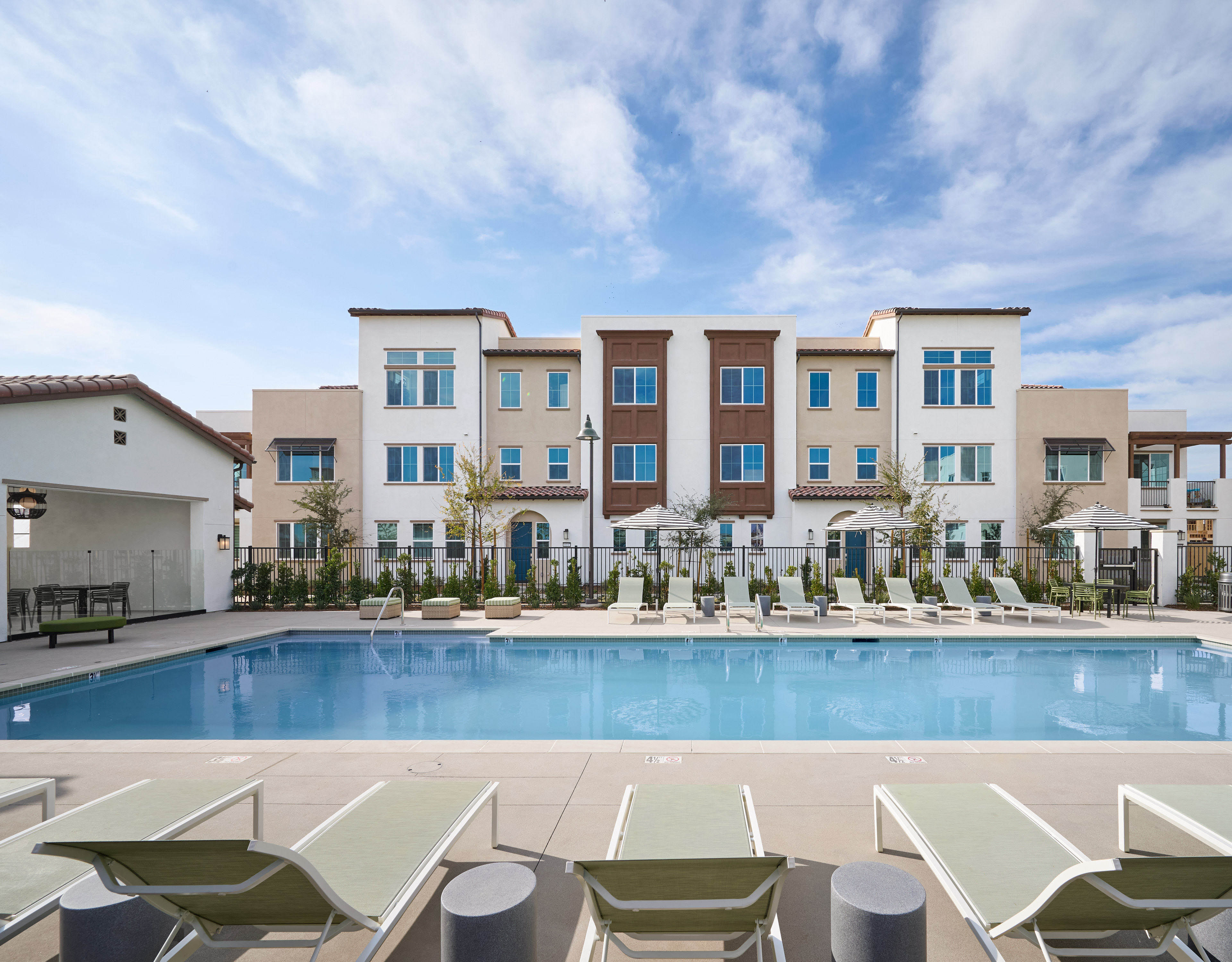 A modern multi-story apartment building with a swimming pool in the foreground, surrounded by lounge chairs and a lush outdoor environment under a bright, cloudy sky.