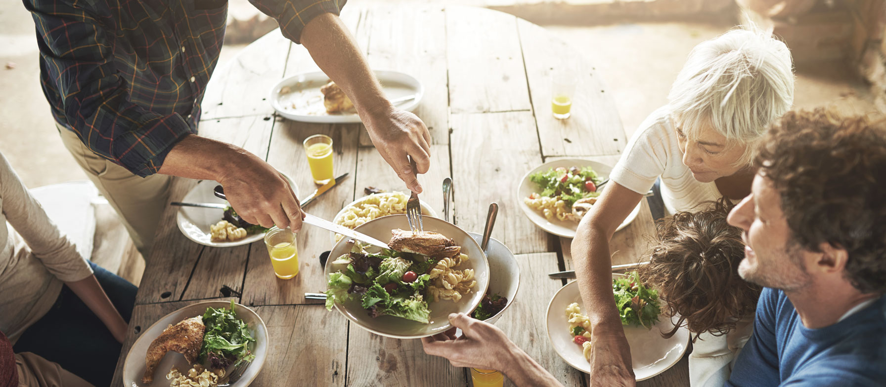 The image depicts a group of people gathered around a wooden table, enjoying a meal together. The foreground shows various plates of food, including salads and sandwiches, while the background suggests a casual, outdoor setting.