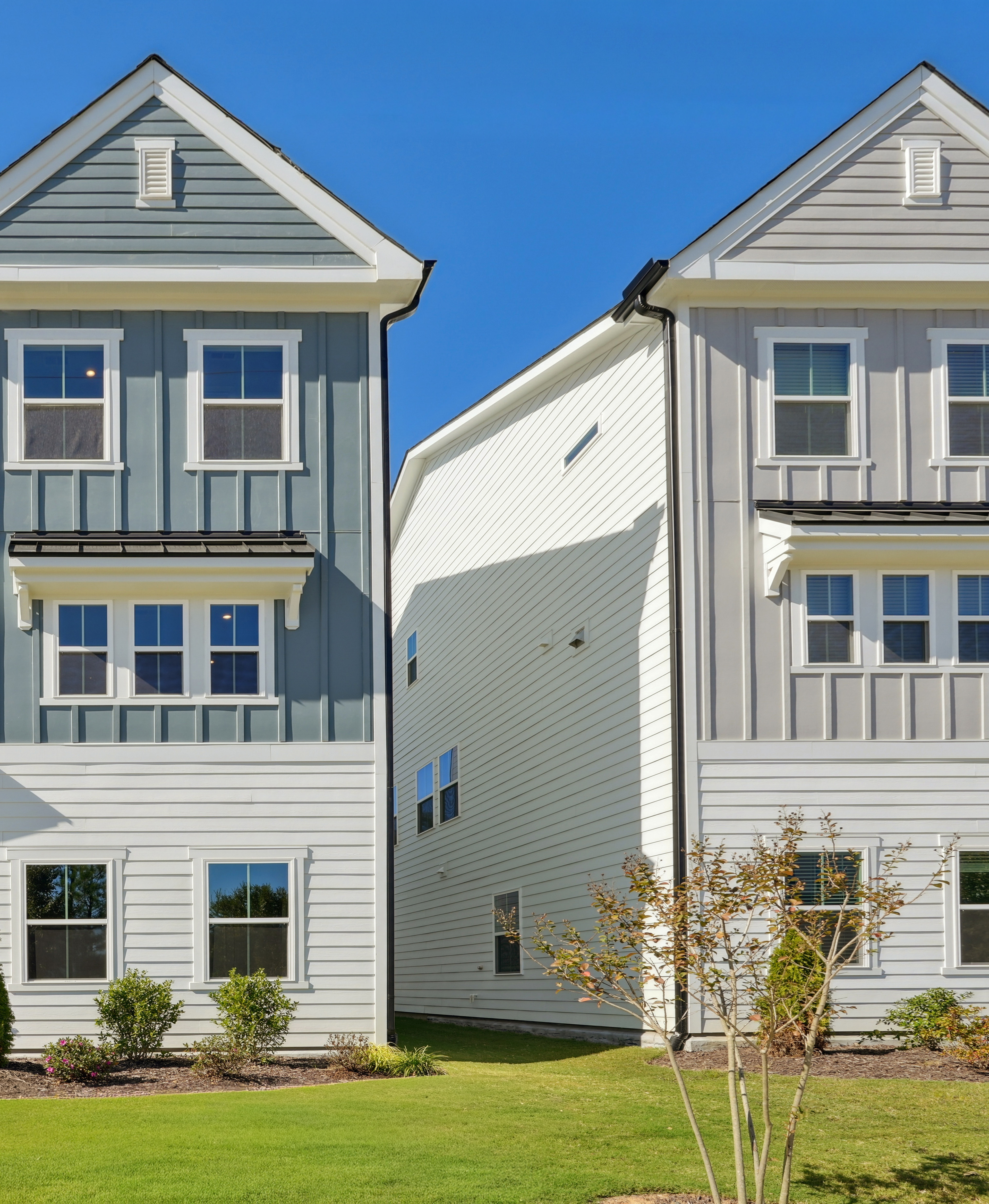 A row of modern, two-story townhouses with gray siding, white trim, and gabled roofs set against a clear blue sky, surrounded by lush greenery in the foreground.