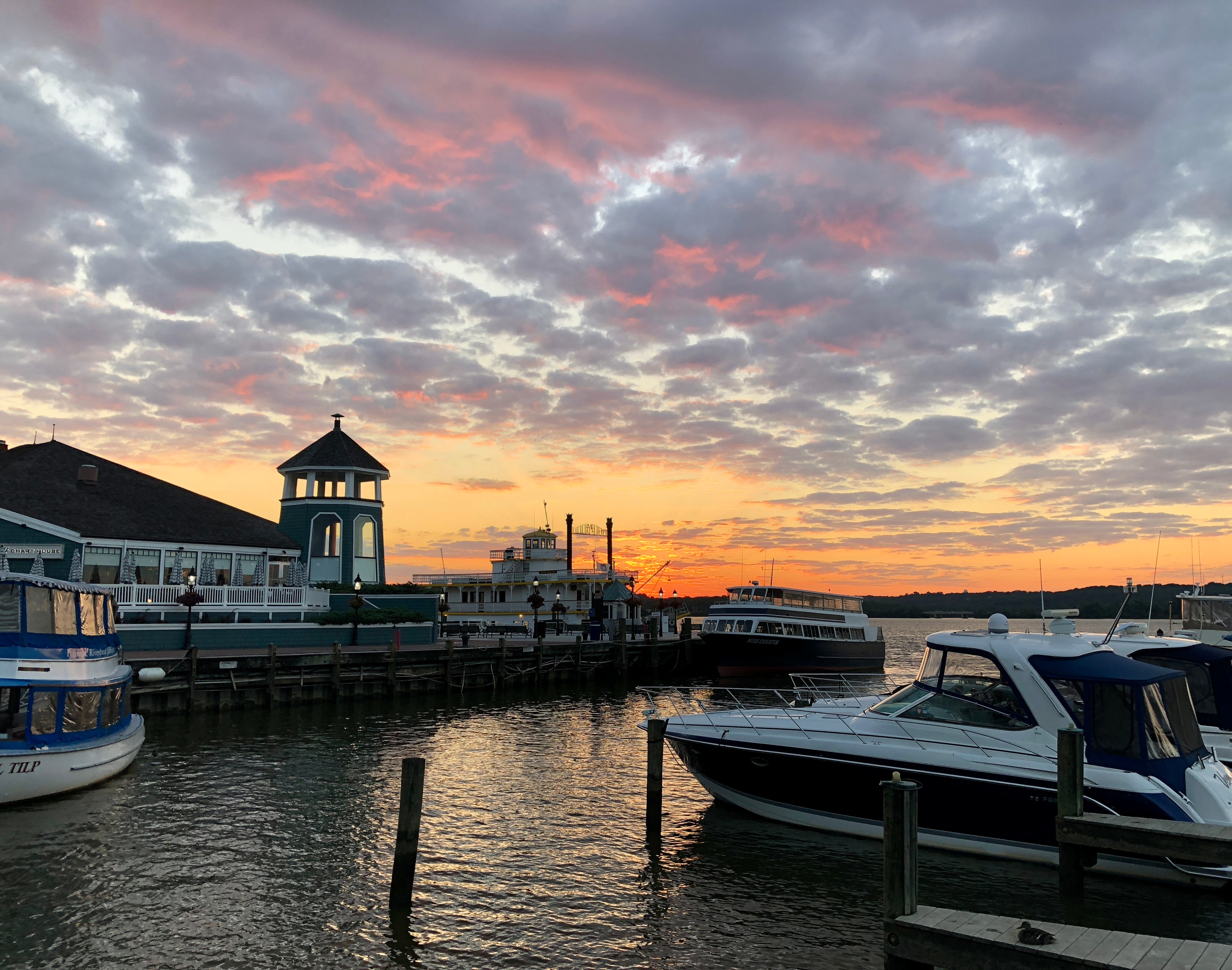 A picturesque harbor scene with boats docked along the waterfront, set against a dramatic sky filled with vibrant hues of orange and pink at sunset.