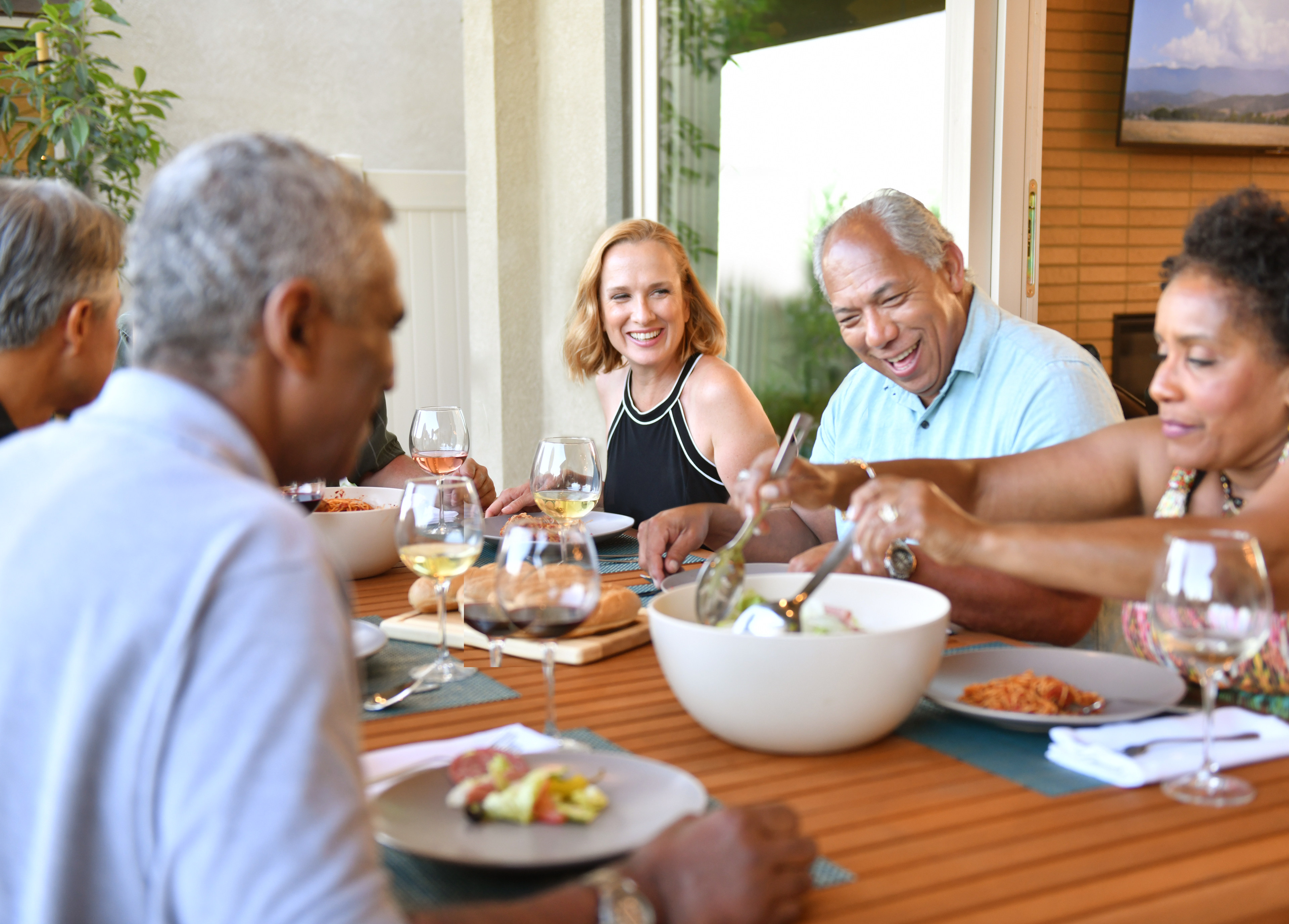 A group of people enjoying a meal together outdoors on a wooden deck, surrounded by lush greenery and a scenic background.