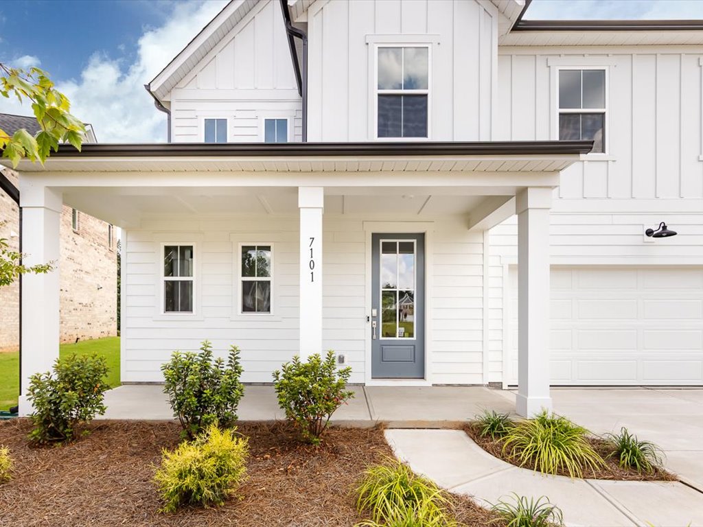 A well-maintained white farmhouse-style home with a covered porch, surrounded by lush greenery and a paved walkway leading to the front door.