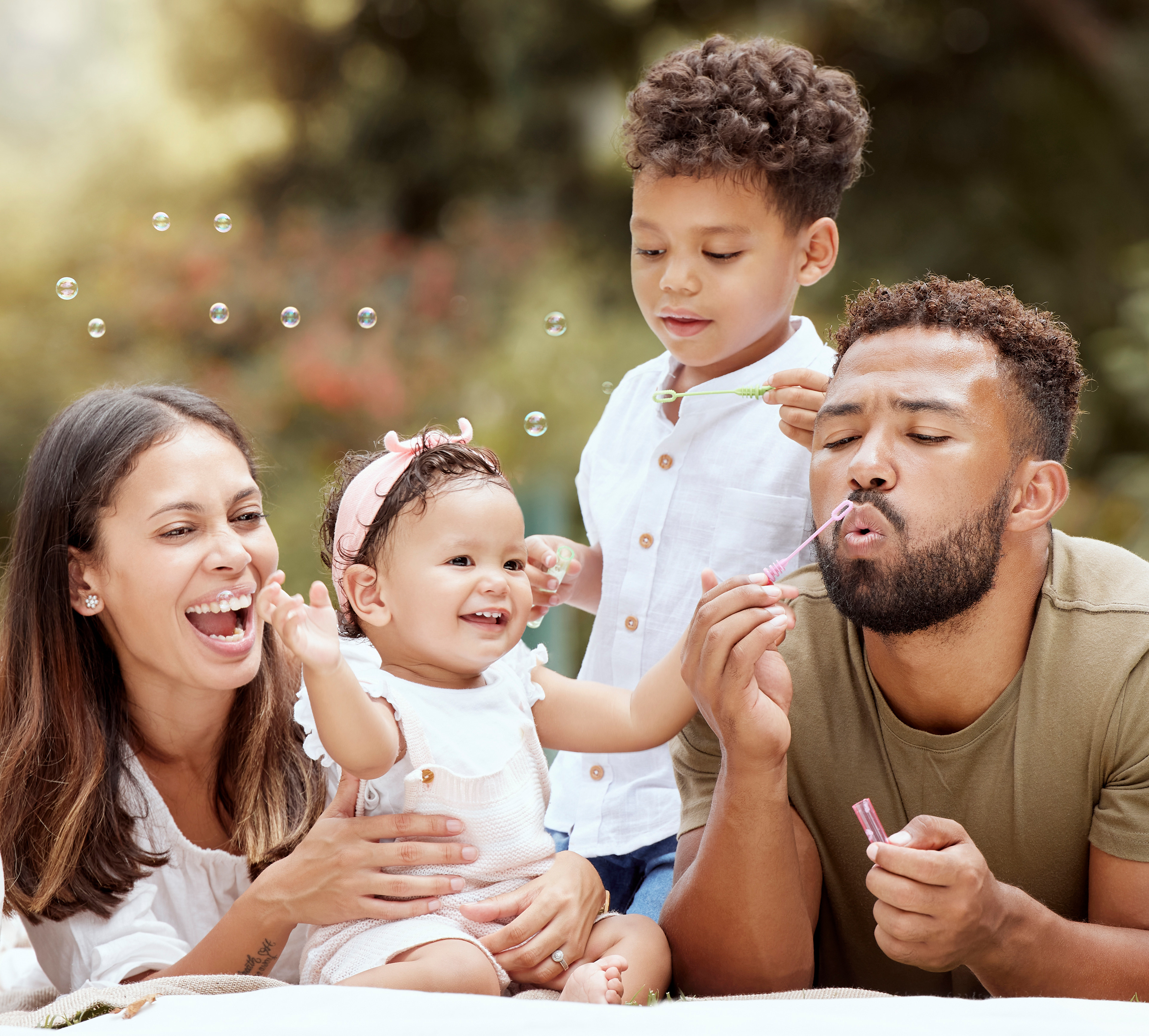 A happy, diverse family sitting together outdoors, surrounded by a blurred natural setting with sunlight filtering through.