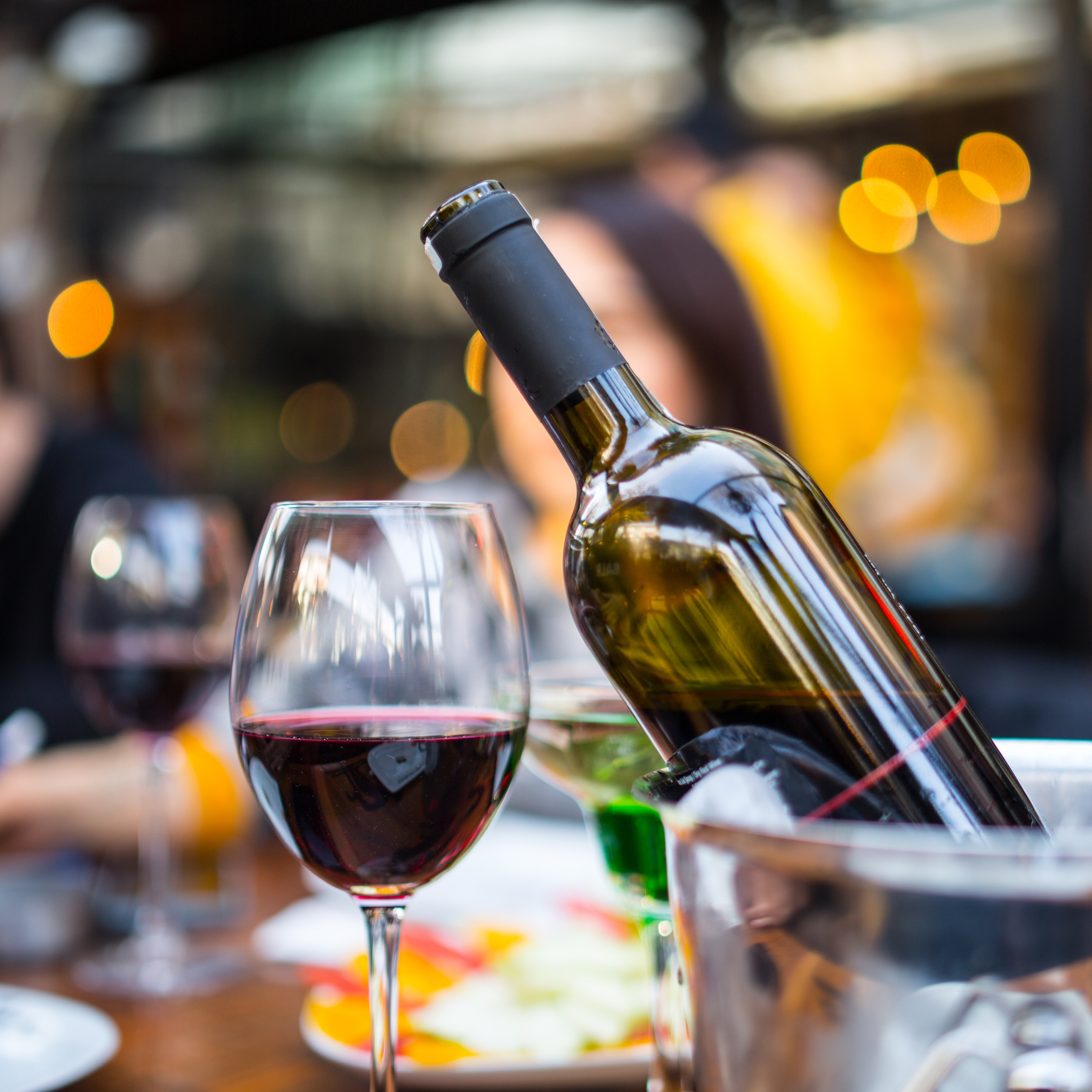 A bottle of wine and a glass of red wine are prominently displayed in the foreground, with a blurred background of what appears to be a restaurant or bar setting with colorful lights.