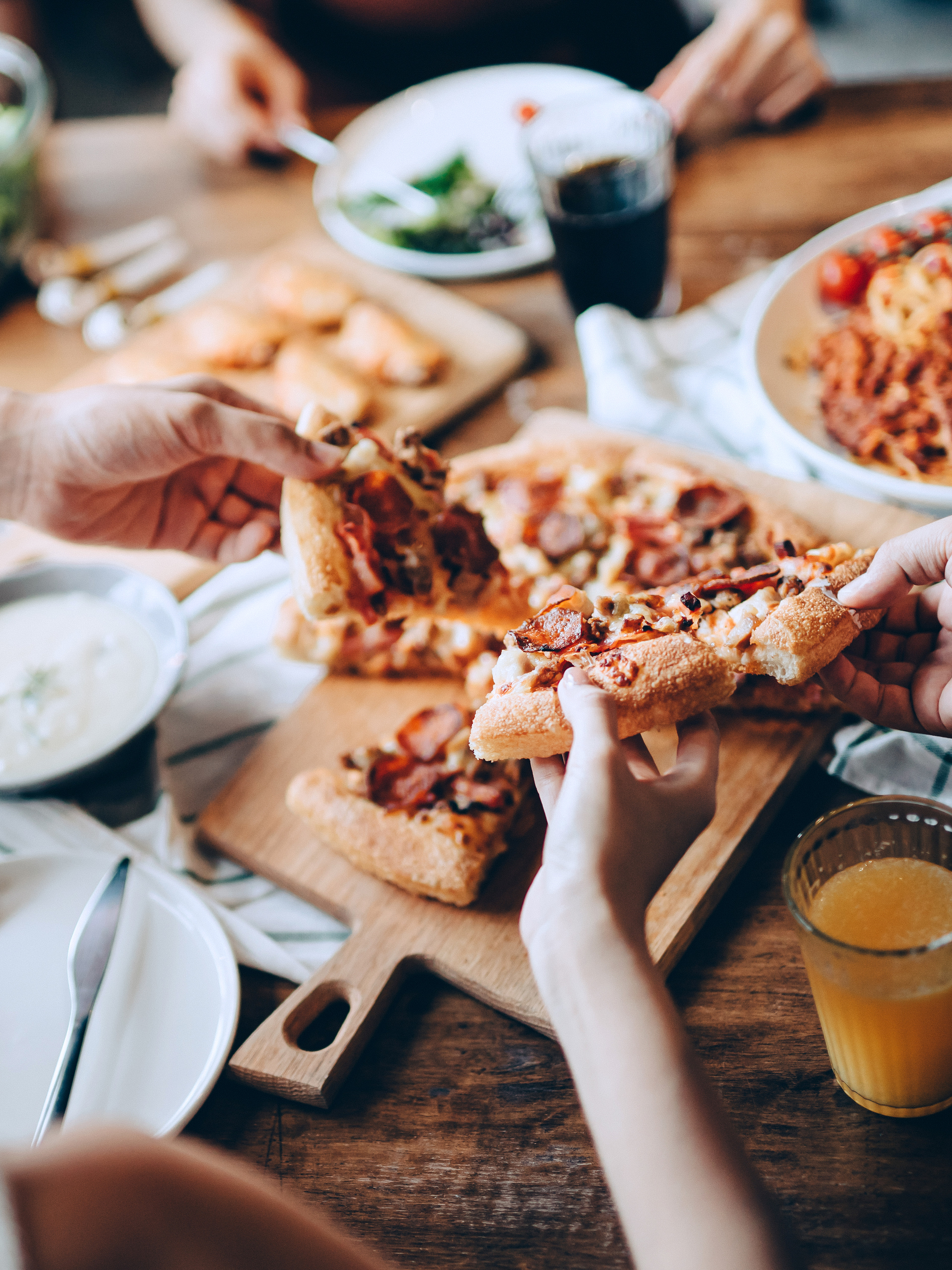 A group of people sharing a meal, with various dishes and drinks spread out on a wooden table in the foreground, while the background appears to be a dimly lit setting.