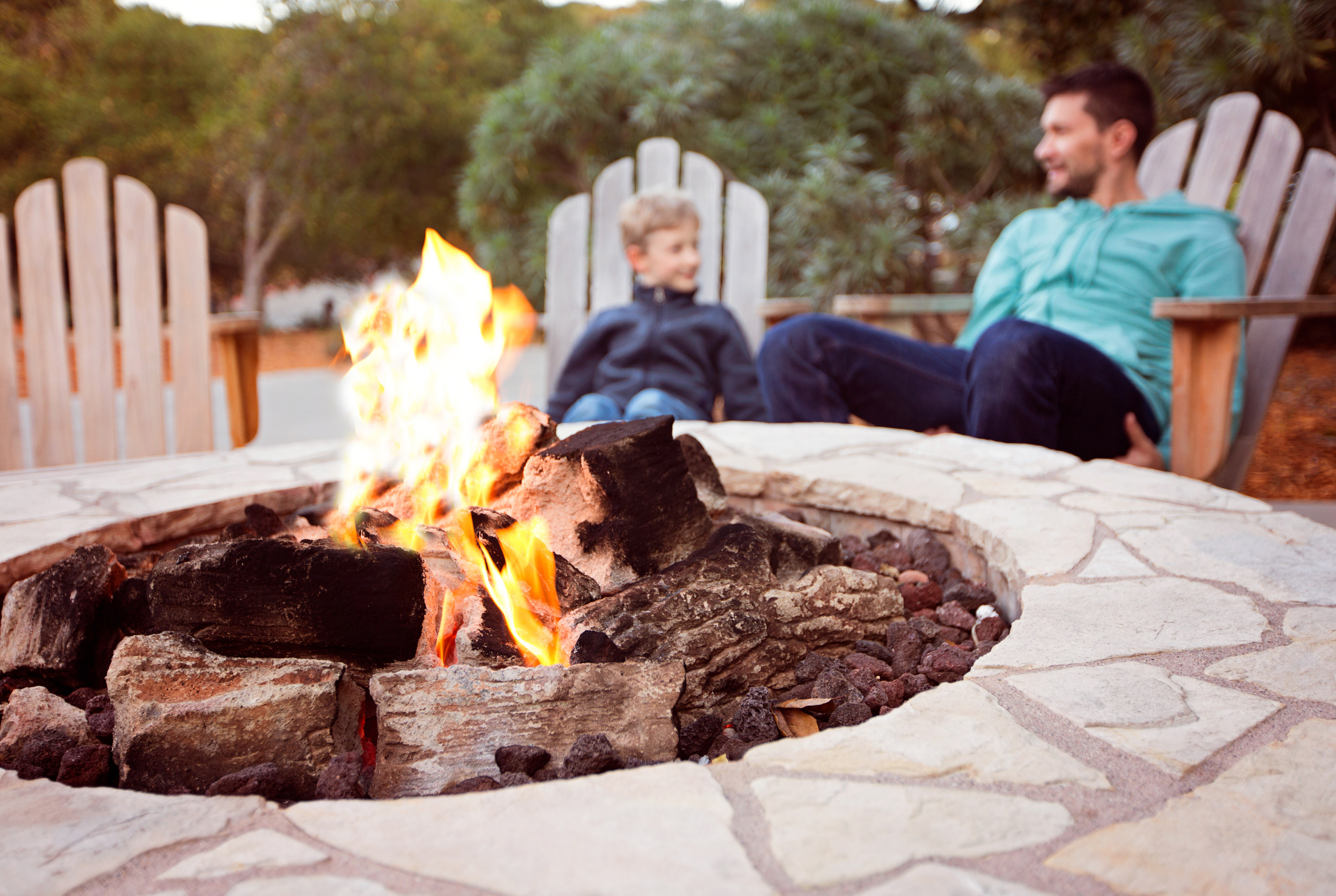 A cozy outdoor fire pit surrounded by stone pavers, with two people sitting on wooden chairs in the background enjoying the warmth and ambiance.