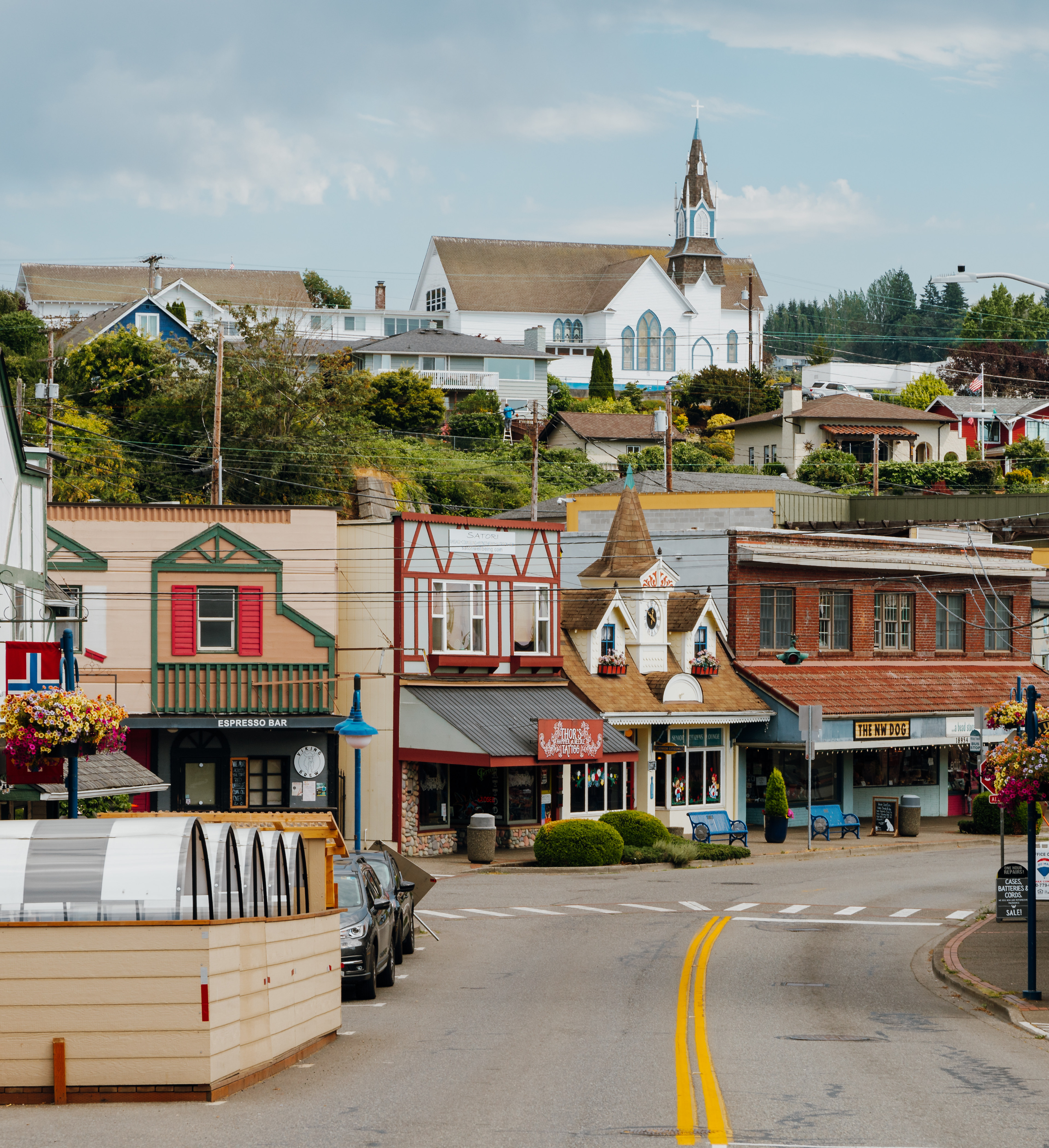 A colorful and quaint town nestled in a hilly landscape, with a mix of traditional and modern buildings, shops, and a church steeple visible in the background.