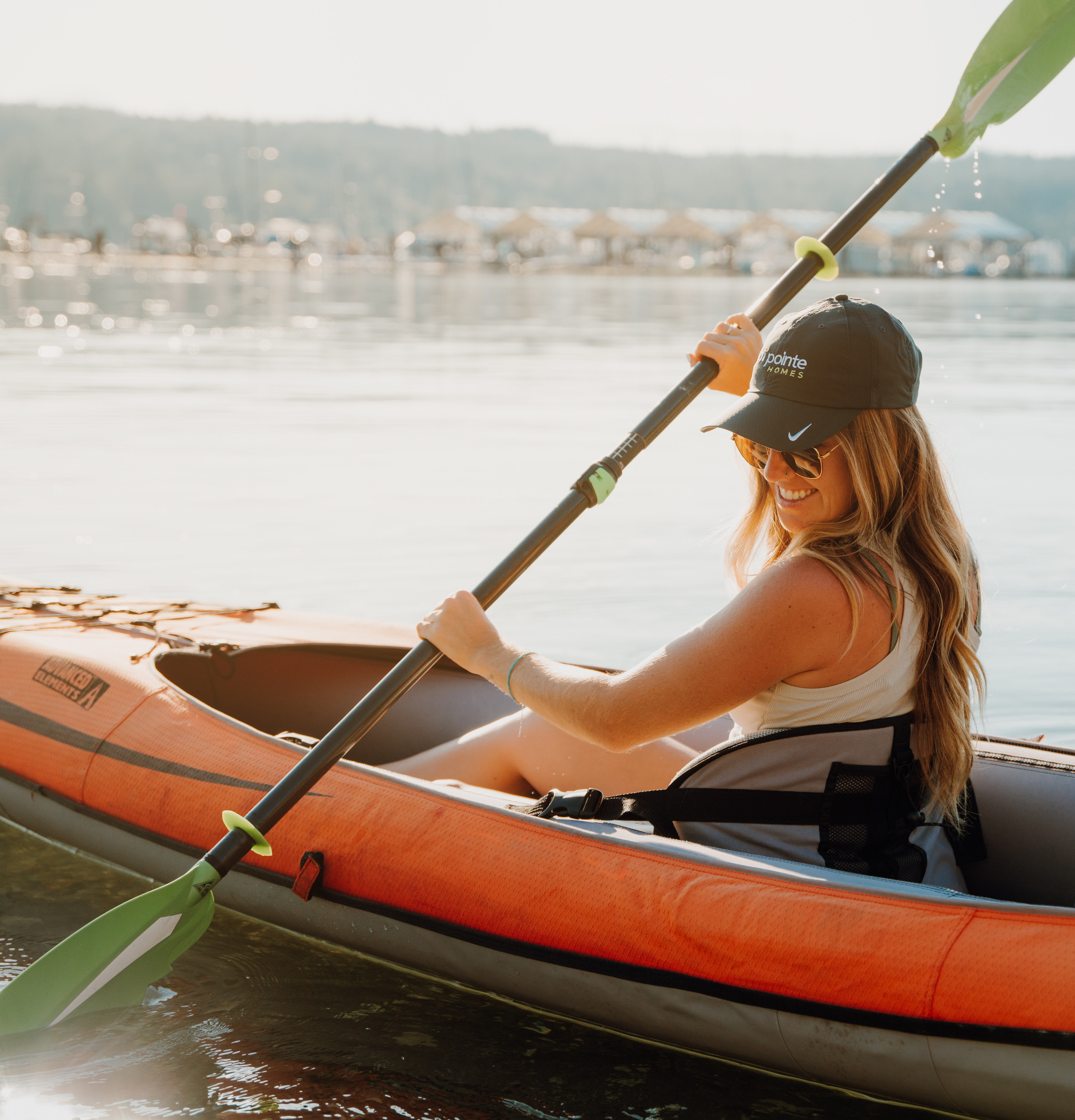 A woman in a bright orange kayak paddles on a serene lake surrounded by mountains in the background.