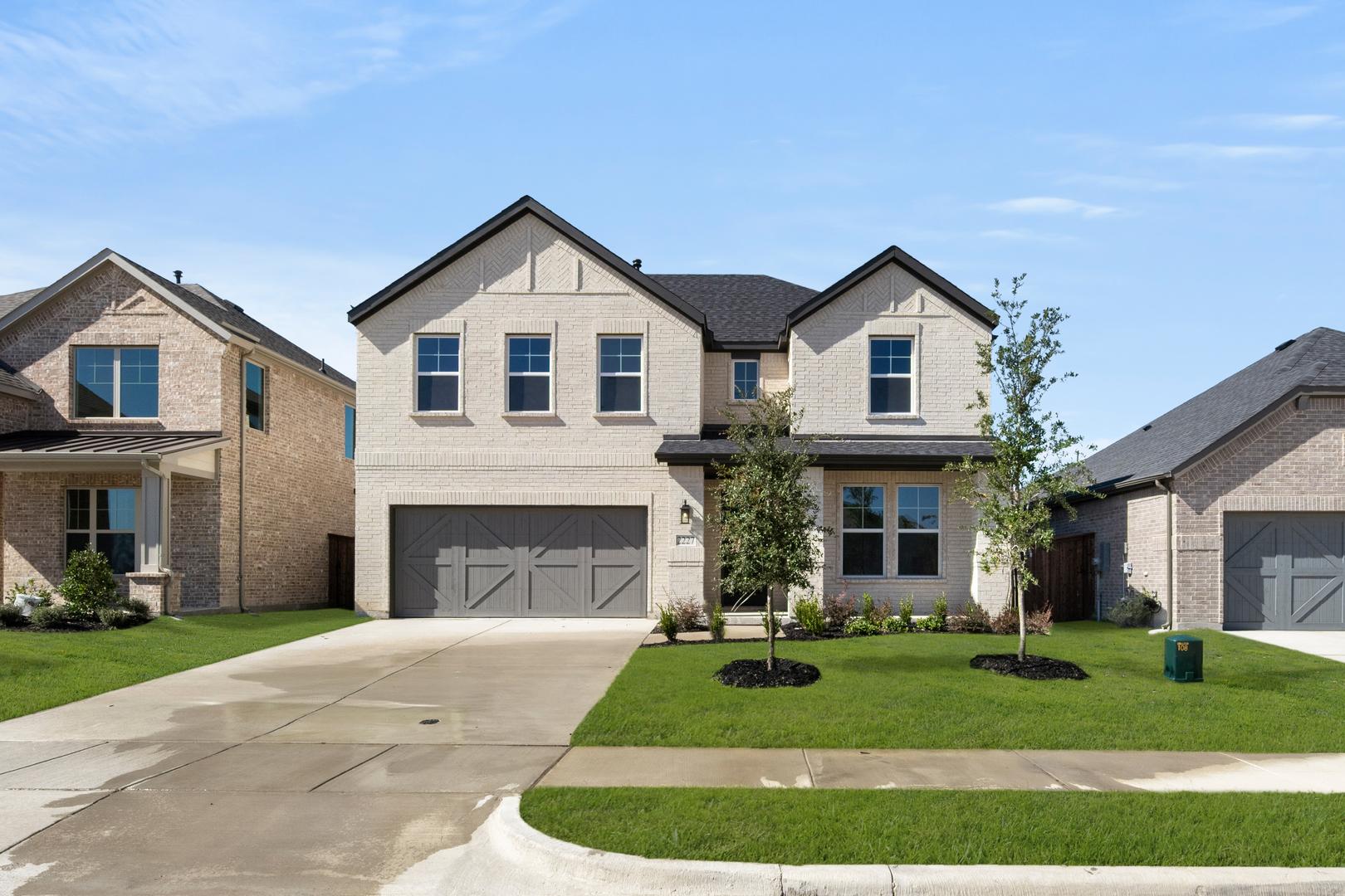A two-story residential house with a garage, surrounded by a well-manicured lawn and other similar houses in the background, set against a clear blue sky.