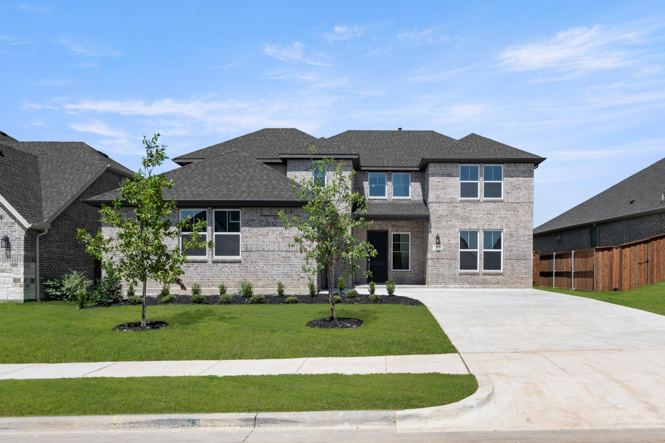 A large, two-story house with a gray exterior and a tiled roof sits on a well-manicured lawn, surrounded by trees and a paved driveway leading to the front entrance.