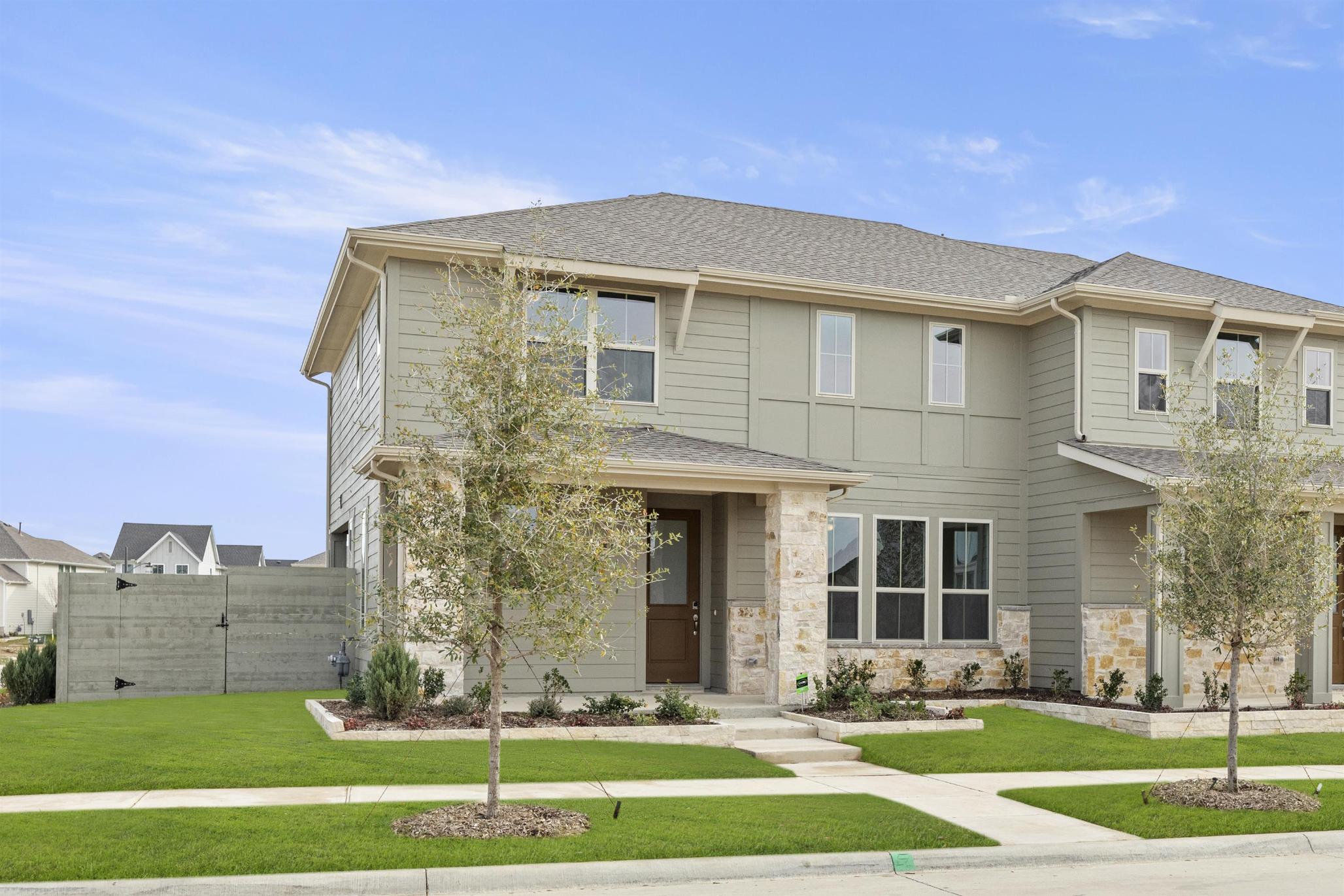 A two-story beige residential building with a well-manicured lawn and landscaping in the foreground, set against a clear blue sky with scattered clouds.