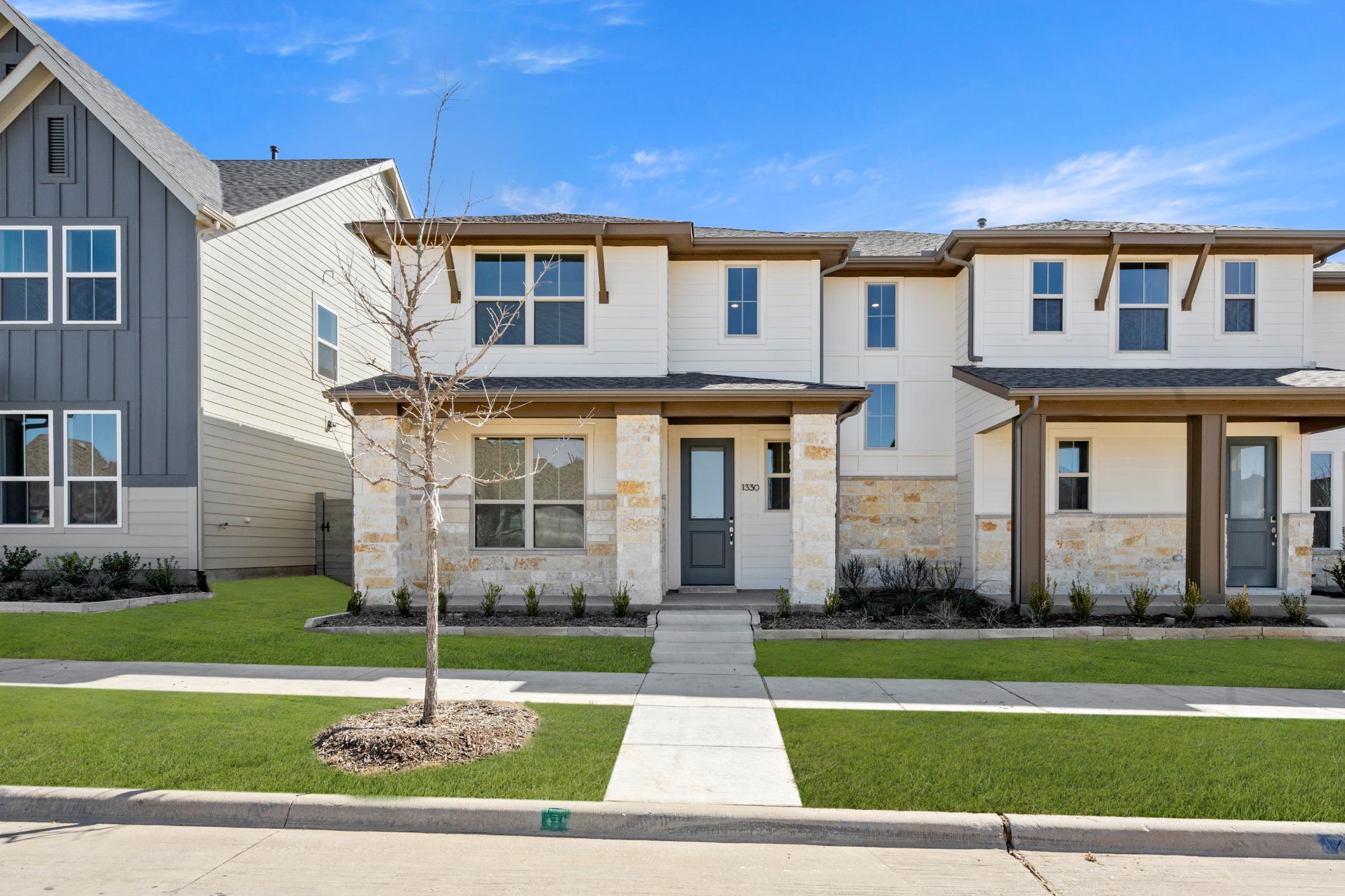 A row of modern, two-story townhouses with gray and white siding, surrounded by a well-manicured lawn and a clear blue sky in the background.